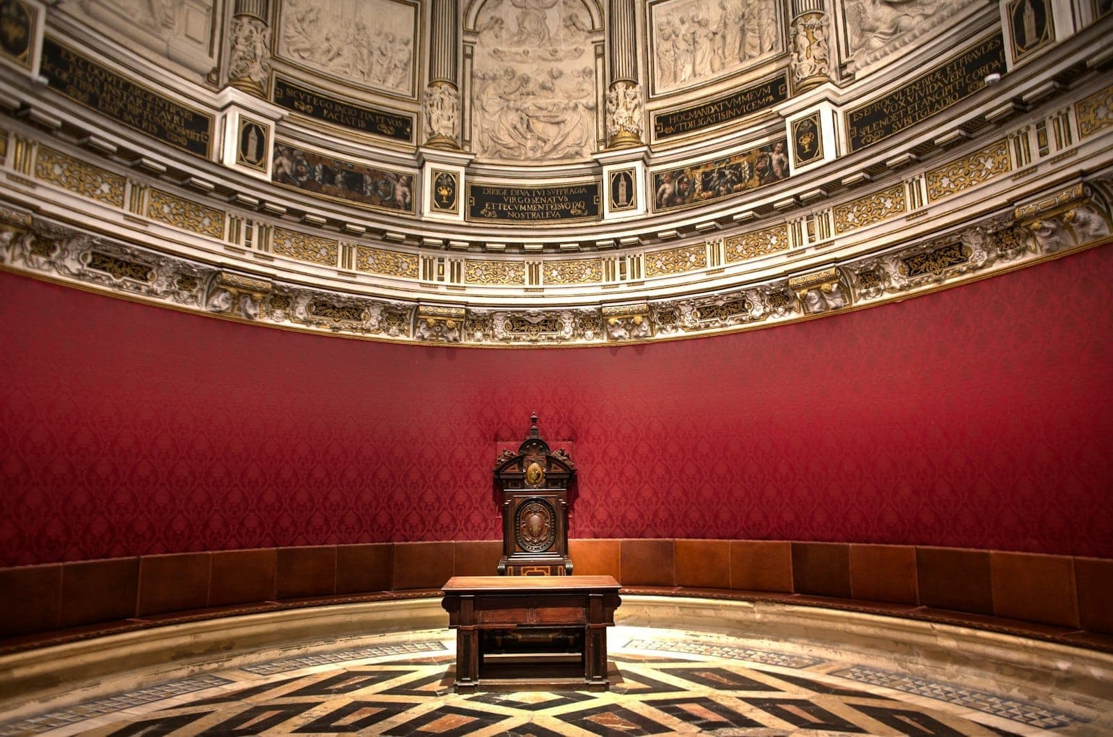 Luxuosa sala do trono circular em vermelho e dourado com teto ornamentado e uma única cadeira cerimonial no centro.