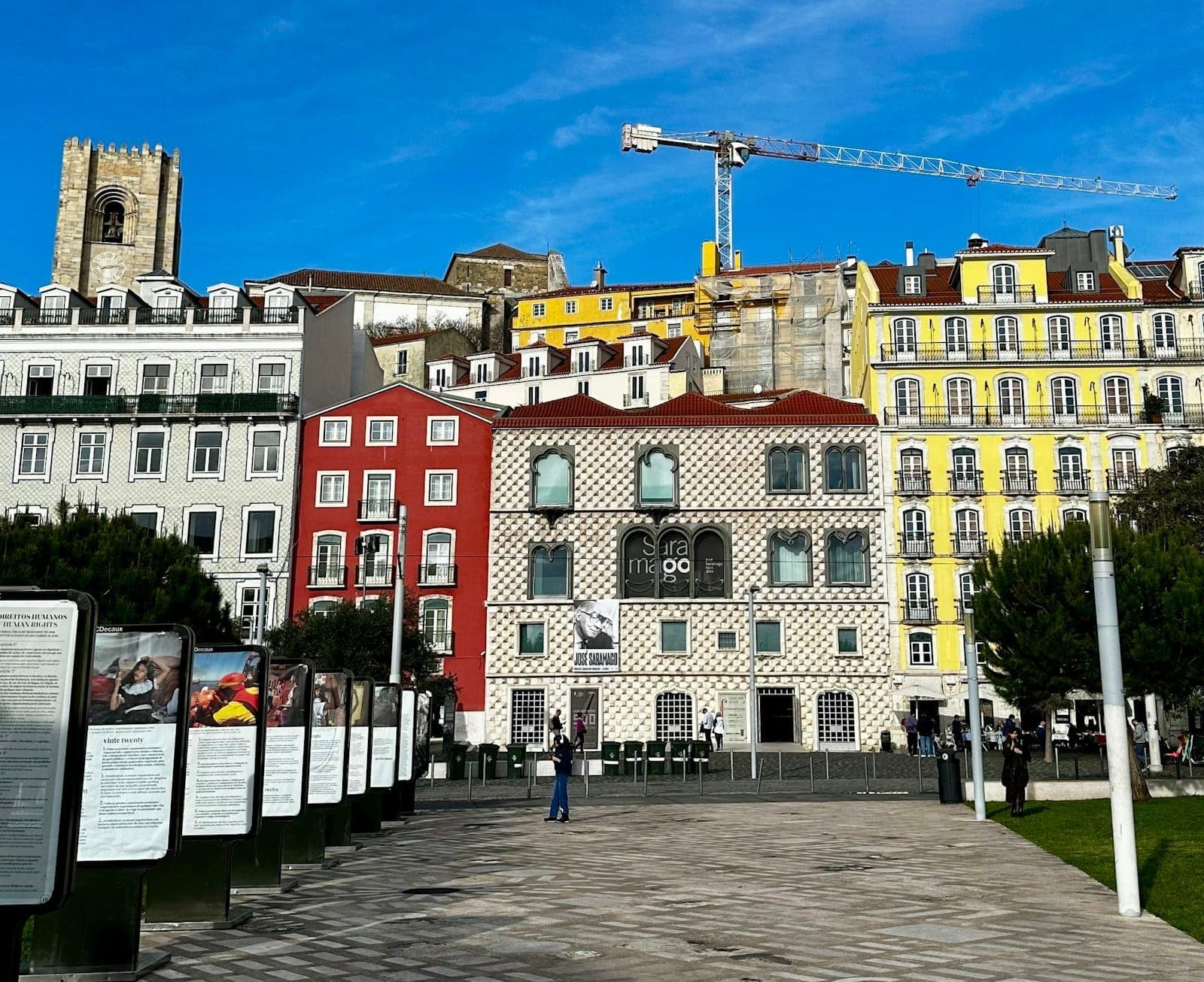 Vista da icônica Casa dos Bicos, sede da Fundação José Saramago, rodeada de coloridos edifícios históricos sob um céu azul, com painéis informativos em primeiro plano.