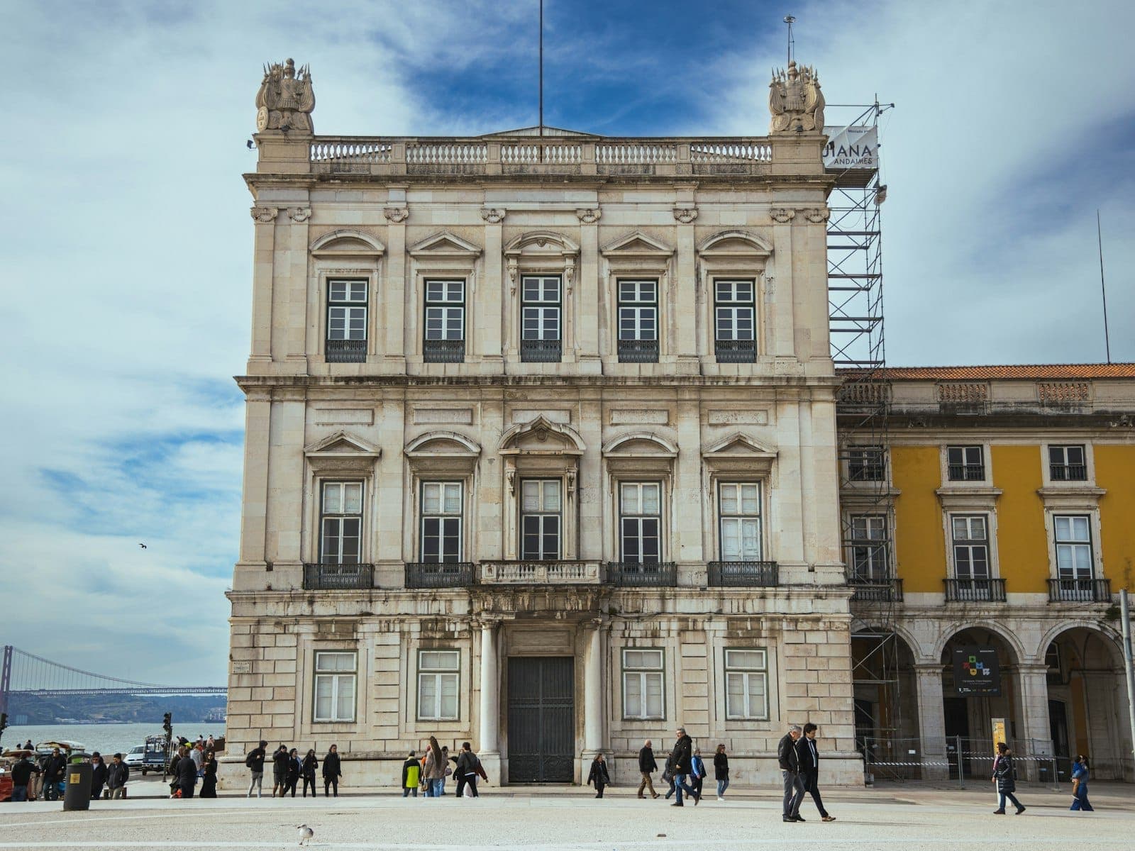 Edifício neoclássico ornamentado na Praça do Comércio de Lisboa, pessoas caminhando em frente, céu azul e vista parcial de um edifício amarelo ao lado.