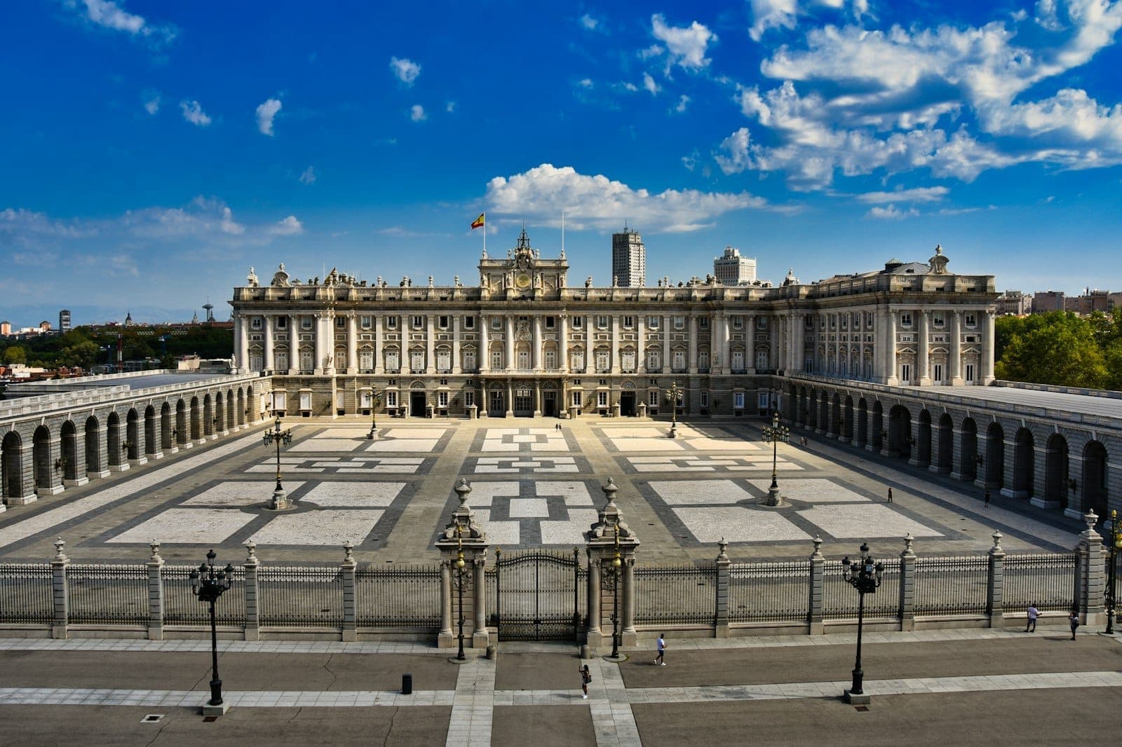 Szeroki widok na dziedziniec Palacio Real w Madrycie pod błękitnym niebem z rozproszonymi chmurami, ukazujący imponującą historyczną architekturę i rozległe place.