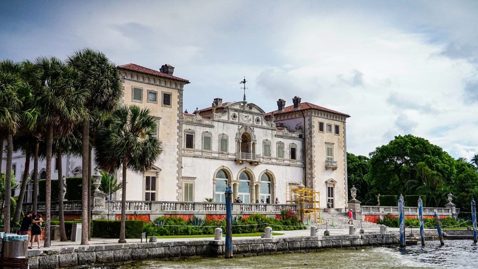 Historic Vizcaya Museum and Gardens mansion in Miami, with palm trees and visitors along the waterfront under a cloudy sky.