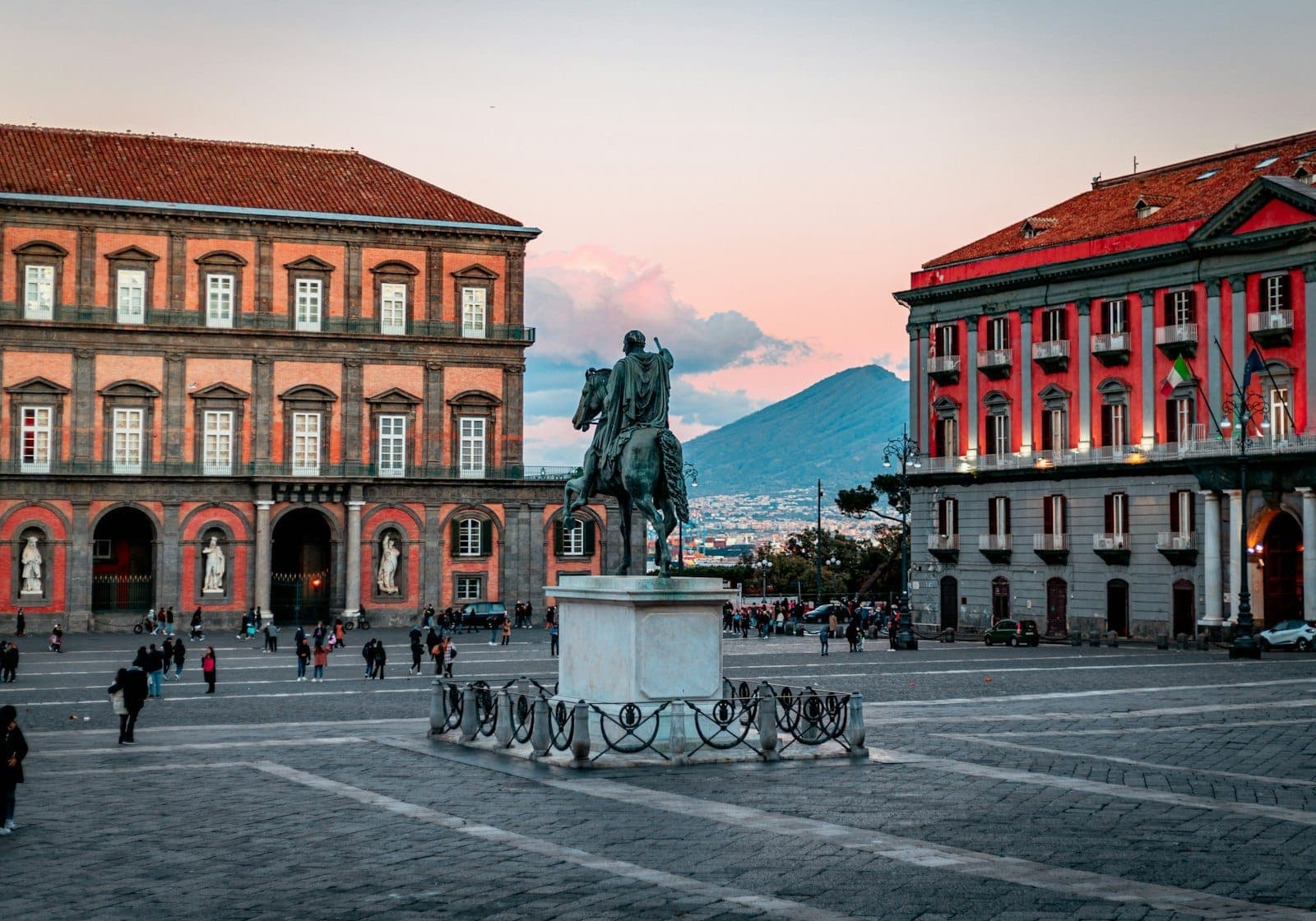 A grandiosa Piazza del Plebiscito de Nápoles ao pôr do sol, com o Palácio Real, estátua equestre e o Monte Vesúvio ao fundo.