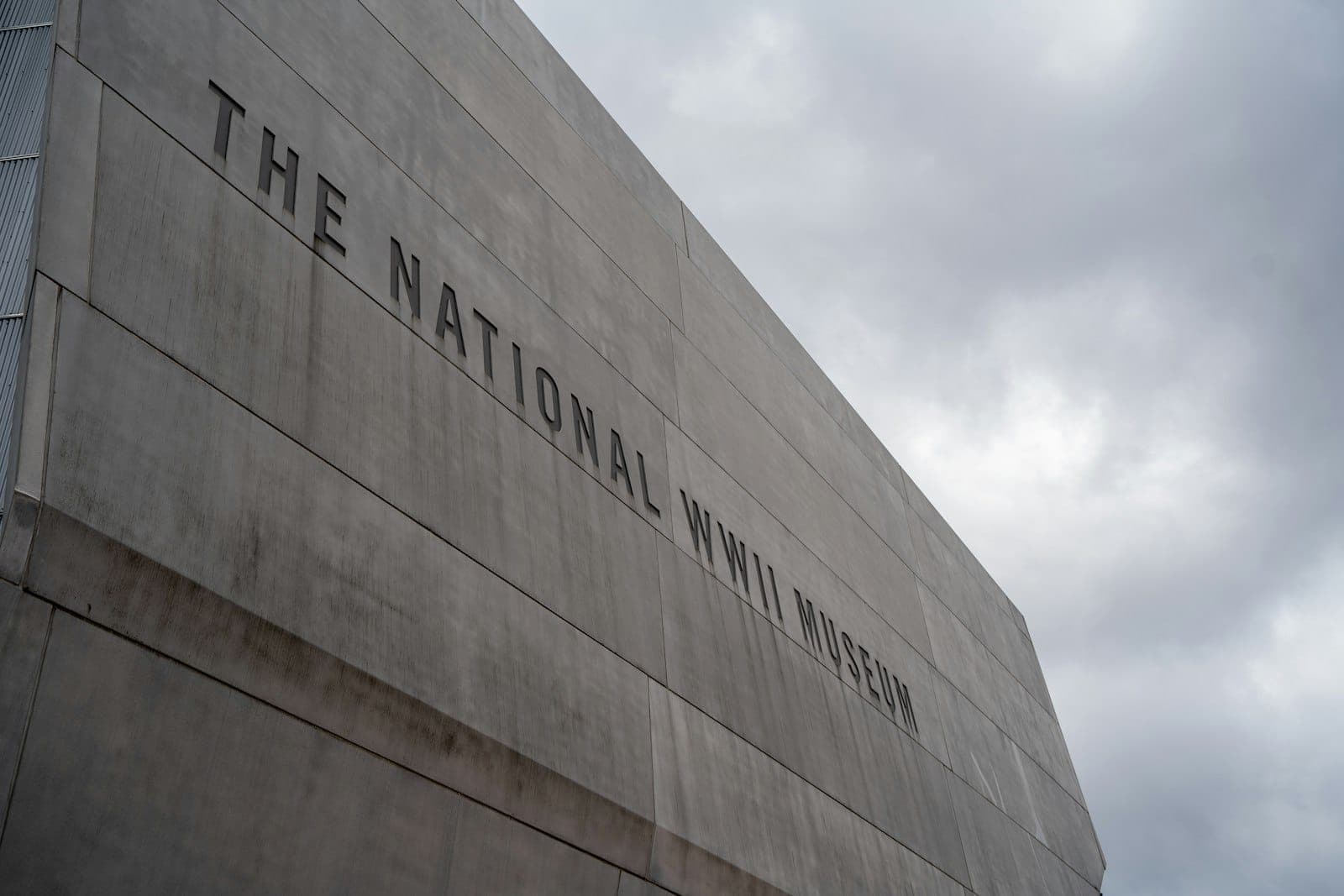 The concrete exterior of The National WWII Museum in New Orleans against a cloudy sky, large lettering visible on the building.