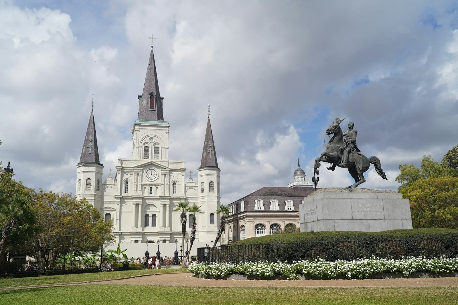 Wide view of St. Louis Cathedral and equestrian statue in Jackson Square, New Orleans, with gardens and clouds overhead.