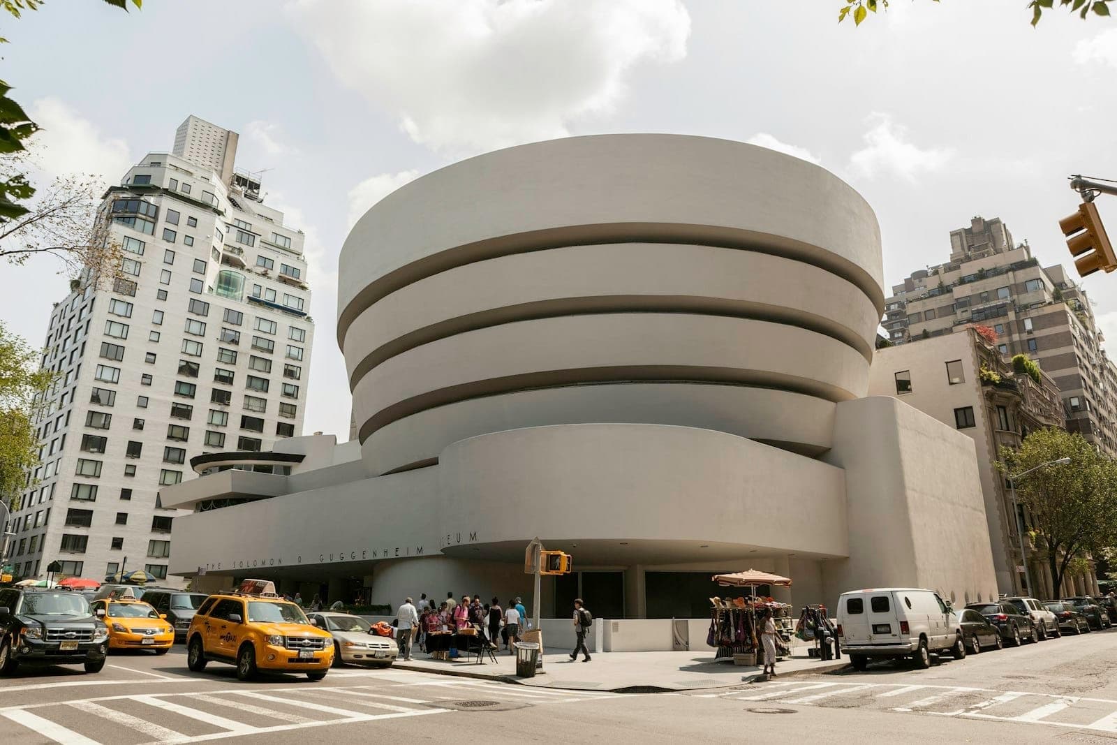 ด้านนอกพิพิธภัณฑ์ Guggenheim ในนิวยอร์ก เห็นตึกโมเดิร์น สัญลักษณ์เมือง พร้อมแท็กซี่สีเหลืองและคนเดินขวักไขว่ที่สี่แยกหน้าพิพิธภัณฑ์