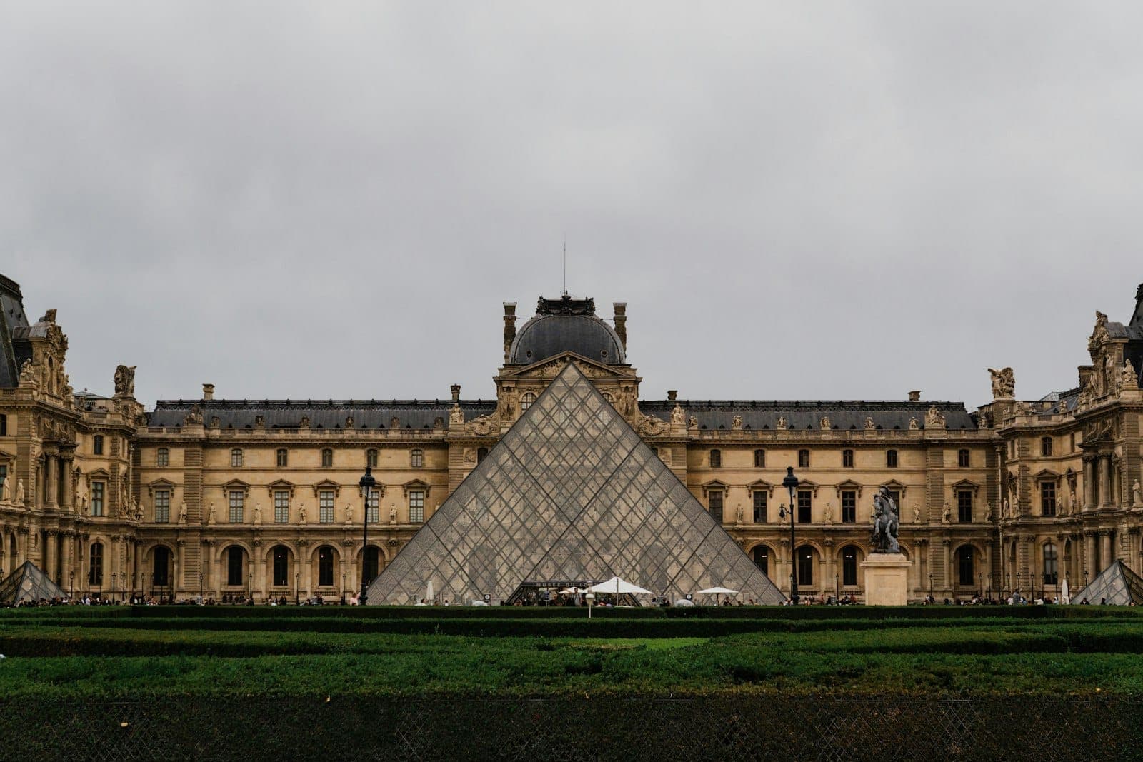 Vue large du Musée du Louvre avec sa pyramide en verre emblématique et sa façade historique sous un ciel nuageux.