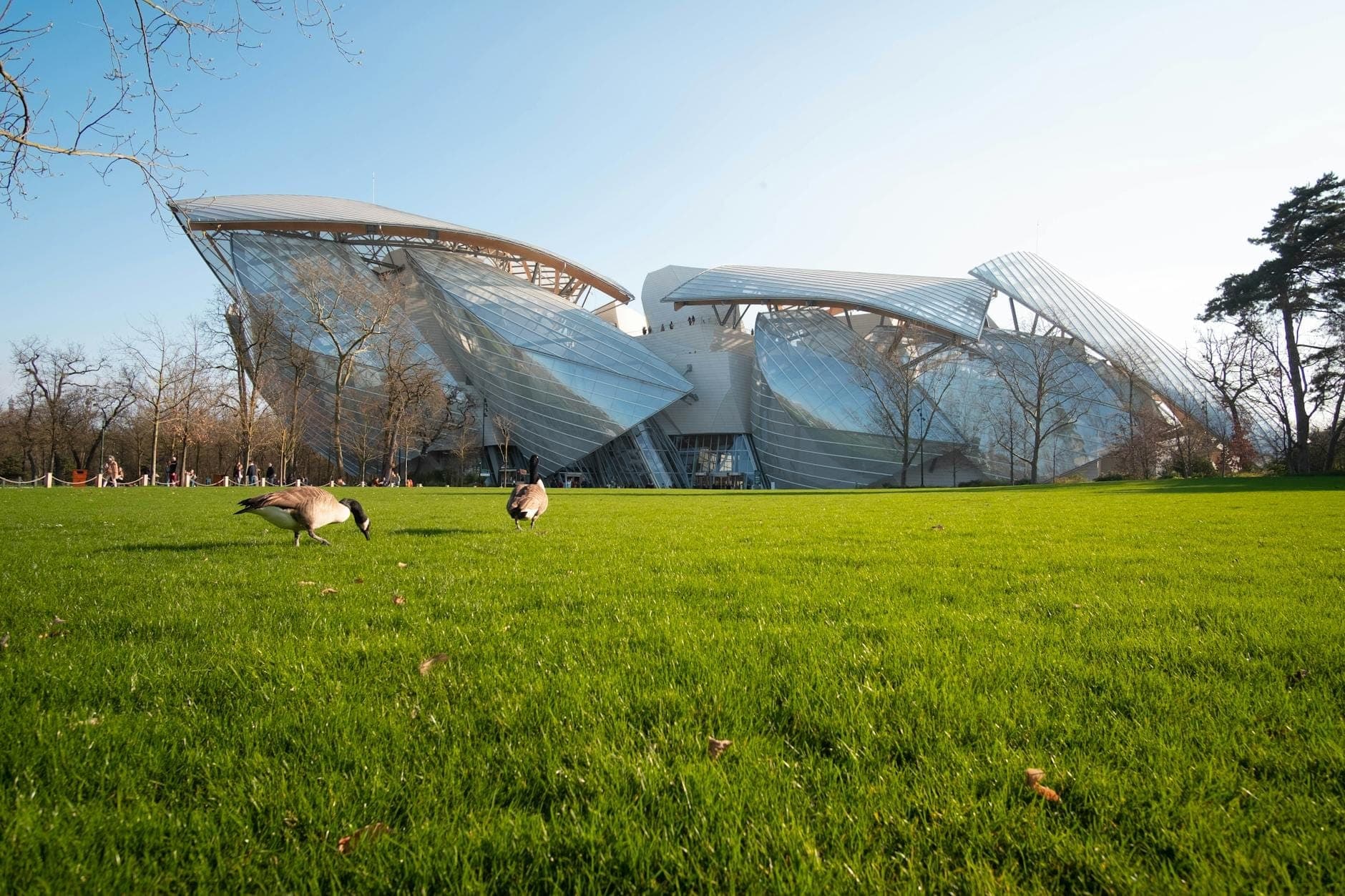 Vue panoramique de l'architecture remarquable en verre et acier de la Fondation Louis Vuitton, vue depuis une pelouse avec des arbres et un ciel bleu.