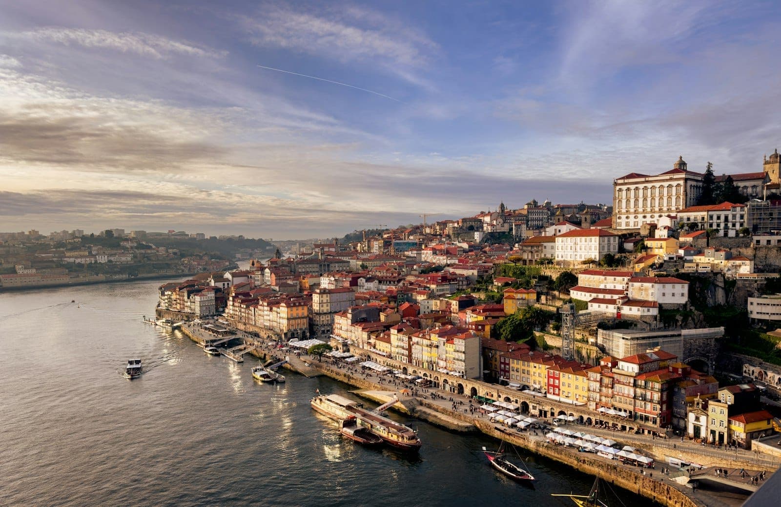Vista panoramica sul colorato lungofiume di Porto con le barche sul Douro e i monumenti della città sotto un cielo drammatico al crepuscolo.