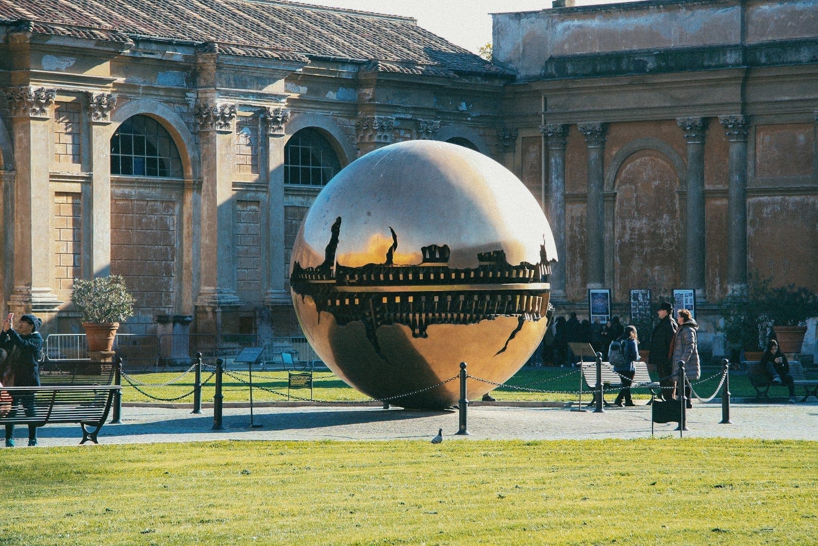Roma'daki çağdaş bir müzenin avlusunda modern bronz küre heykeli (Sphere Within a Sphere), yakınında yürüyen insanlarla birlikte.