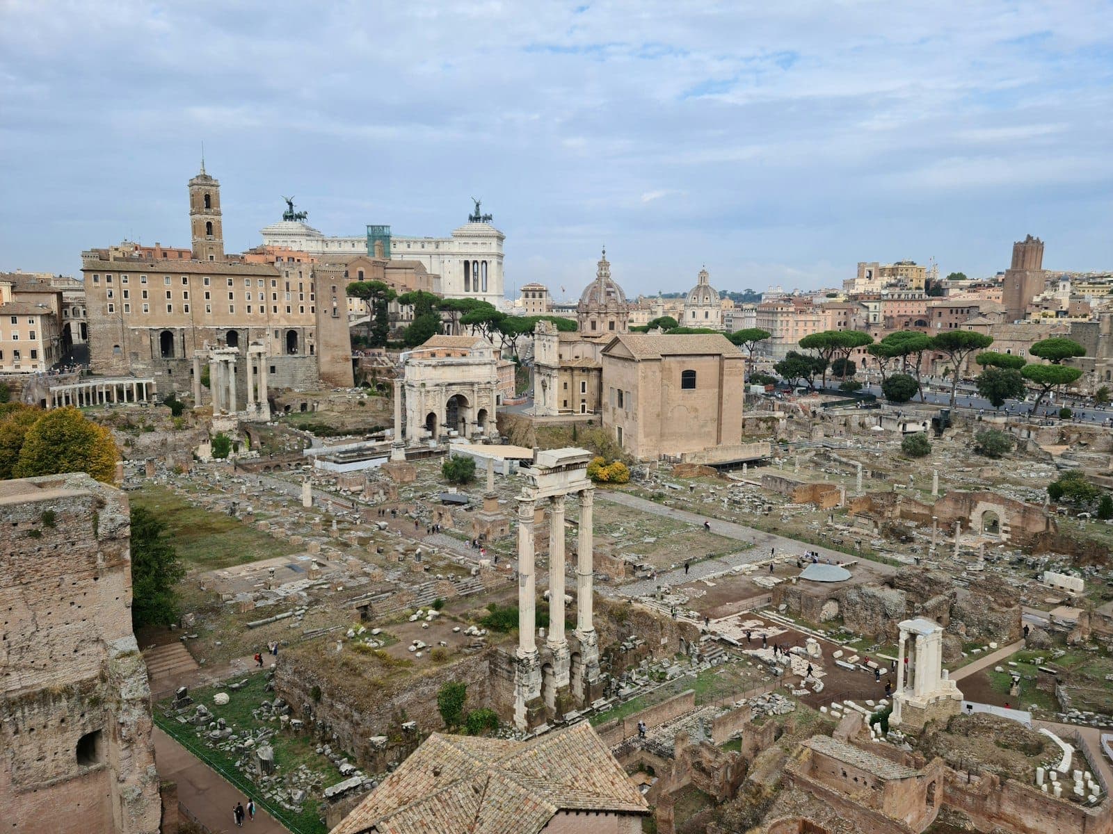 Roma'da mavi bir gökyüzü altında dağınık antik kalıntılar, sütunlar ve tarihi binalarla Roman Forumu'nun geniş görünümü.
