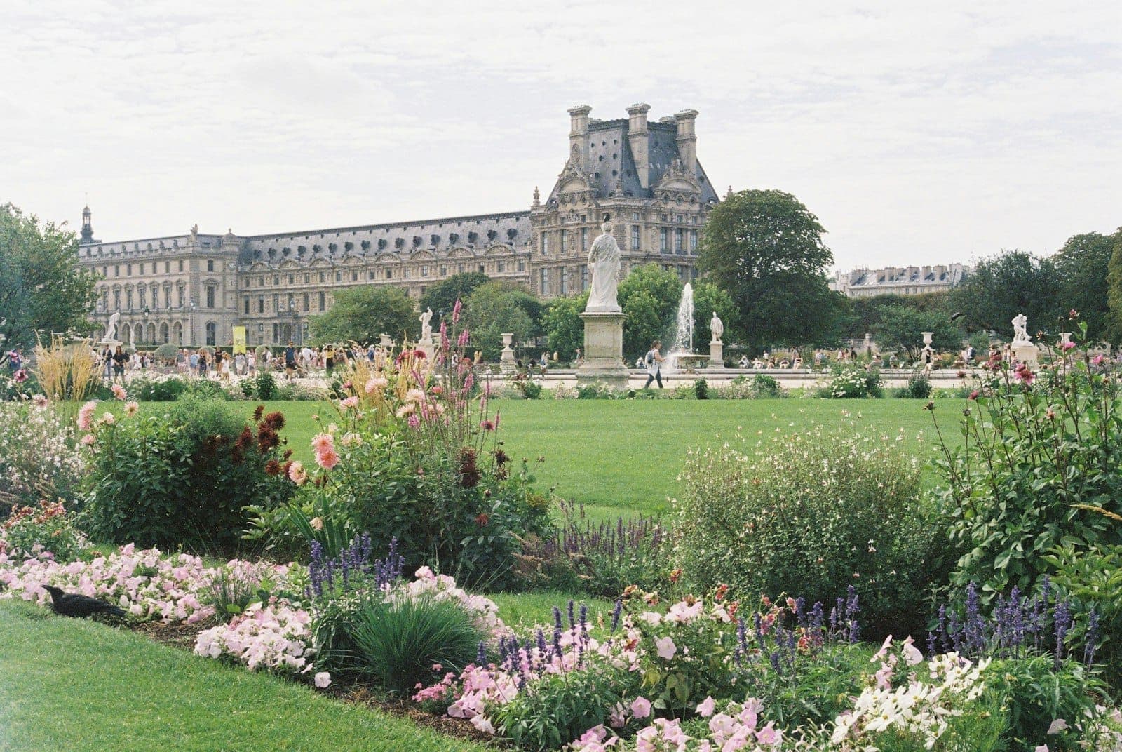 Lush Parisian garden with colorful flowerbeds and statues, classic French architecture in the background under a partly cloudy sky.