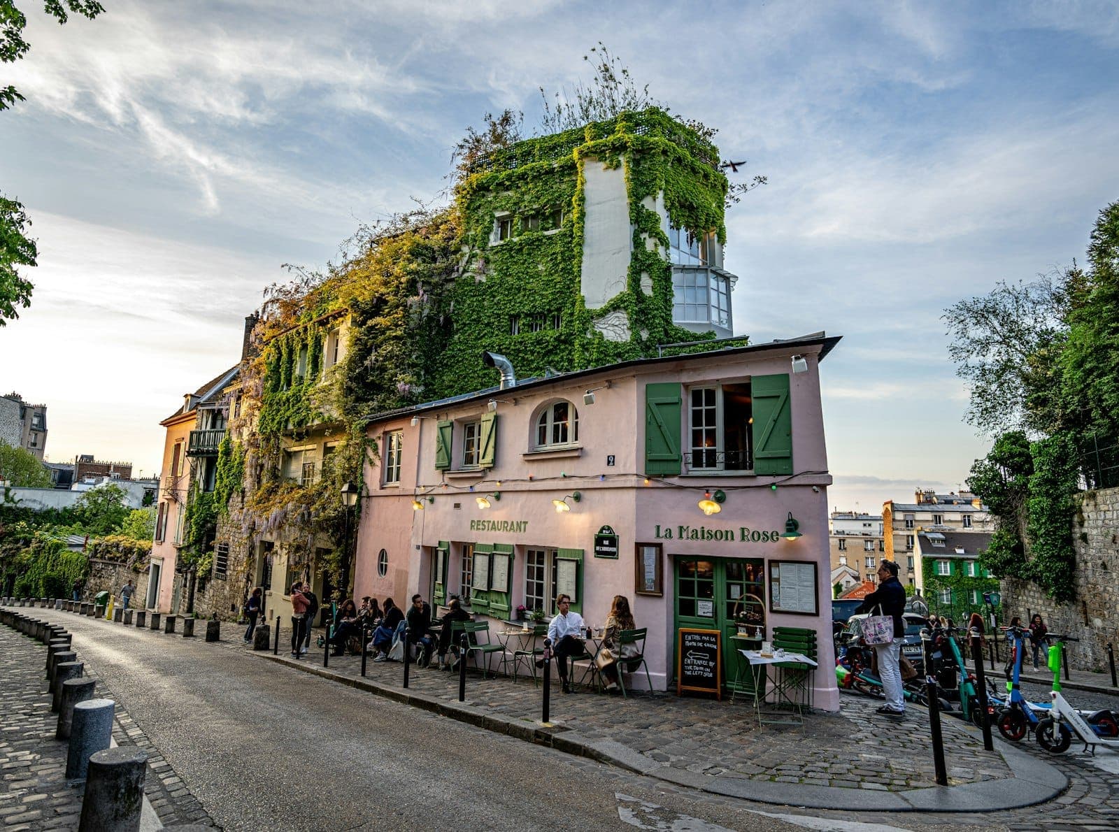 Kafe La Maison Rose yang menawan dan tertutup tanaman ivy di jalan berbatu yang tenang di Montmartre, dengan orang-orang yang makan dan berjalan-jalan saat matahari terbenam.