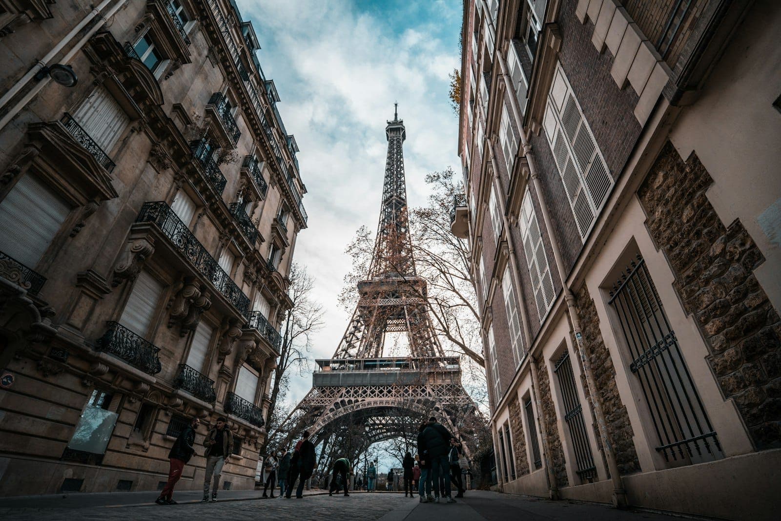 View of the Eiffel Tower framed by classic Parisian apartment buildings, people walking along the street on a cloudy day.