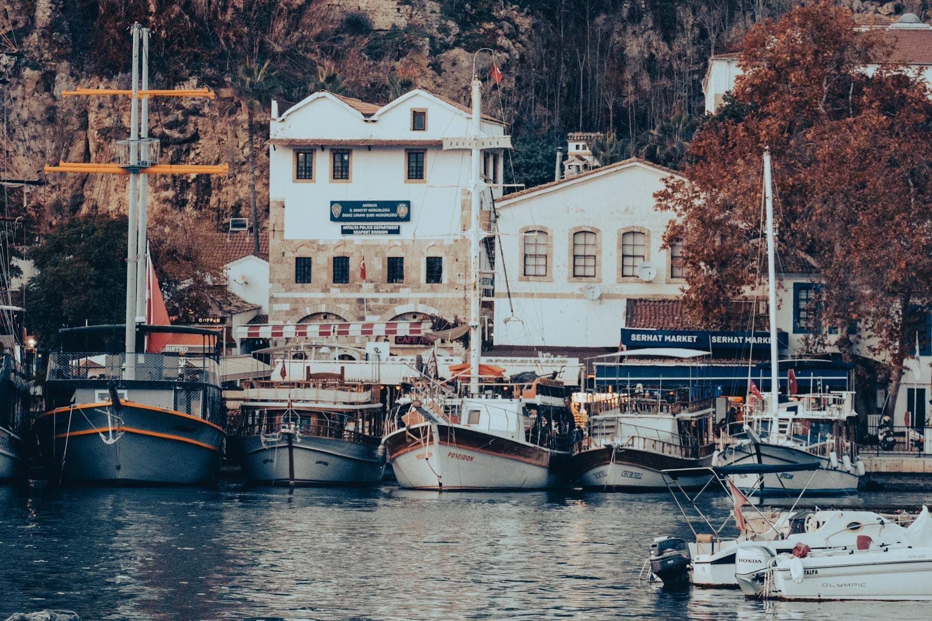 Im alten Hafen von Antalya vertäute Boote mit herbstlich gefärbten Bäumen und historischen Gebäuden im Hintergrund.