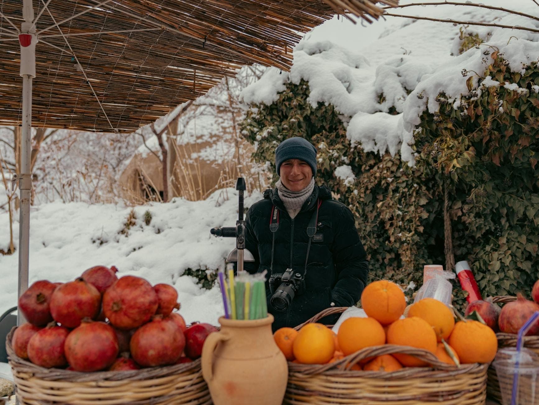 Ein lächelnder Verkäufer steht hinter Körben mit Orangen und Granatäpfeln auf einem Außenmarkt, mit schneebedeckten Büschen und Boden, die winterliche Frische hervorrufen.