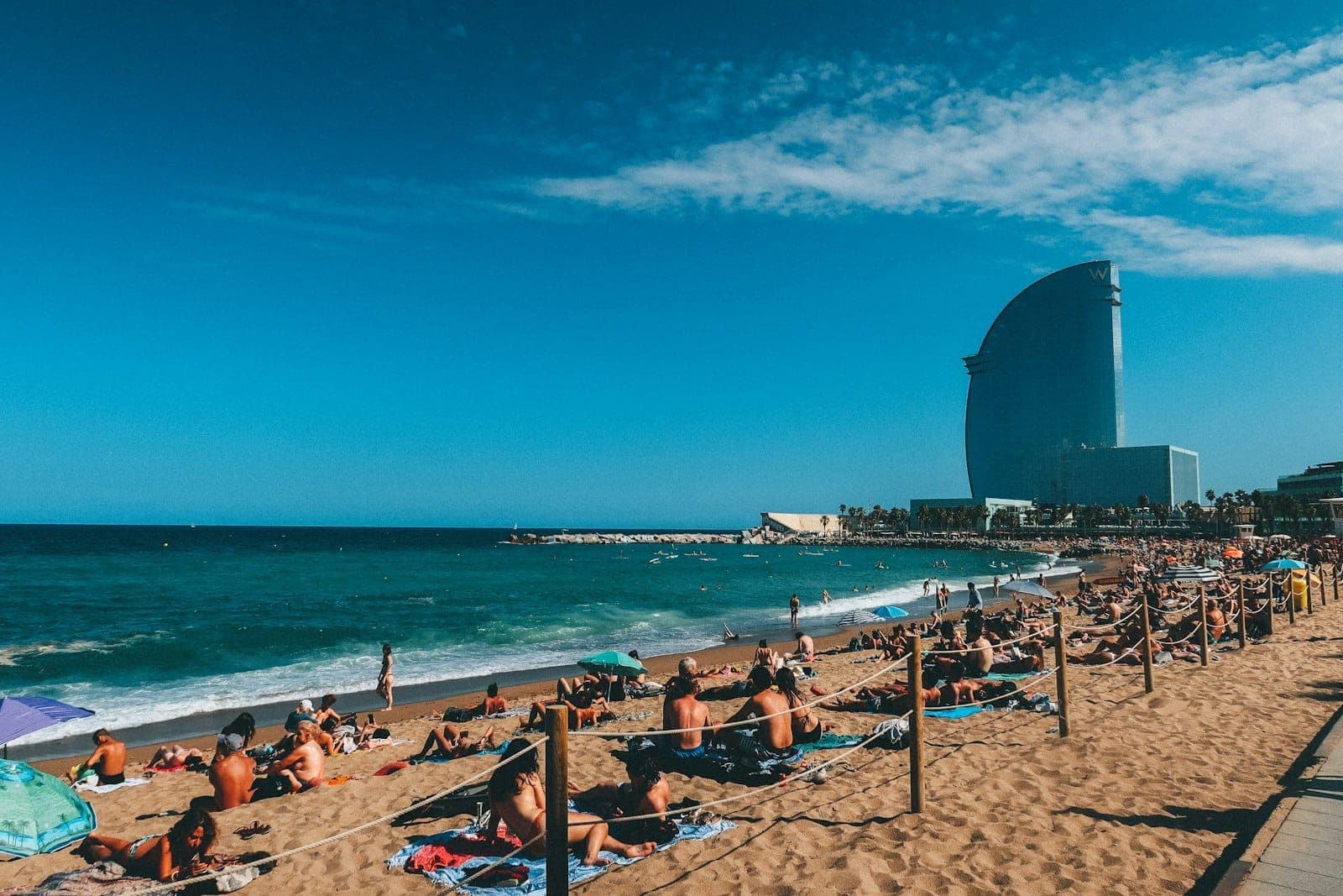 Crowded Barceloneta beach on a sunny day, with people sunbathing and swimming. The iconic W Hotel rises in the background under a clear blue sky.