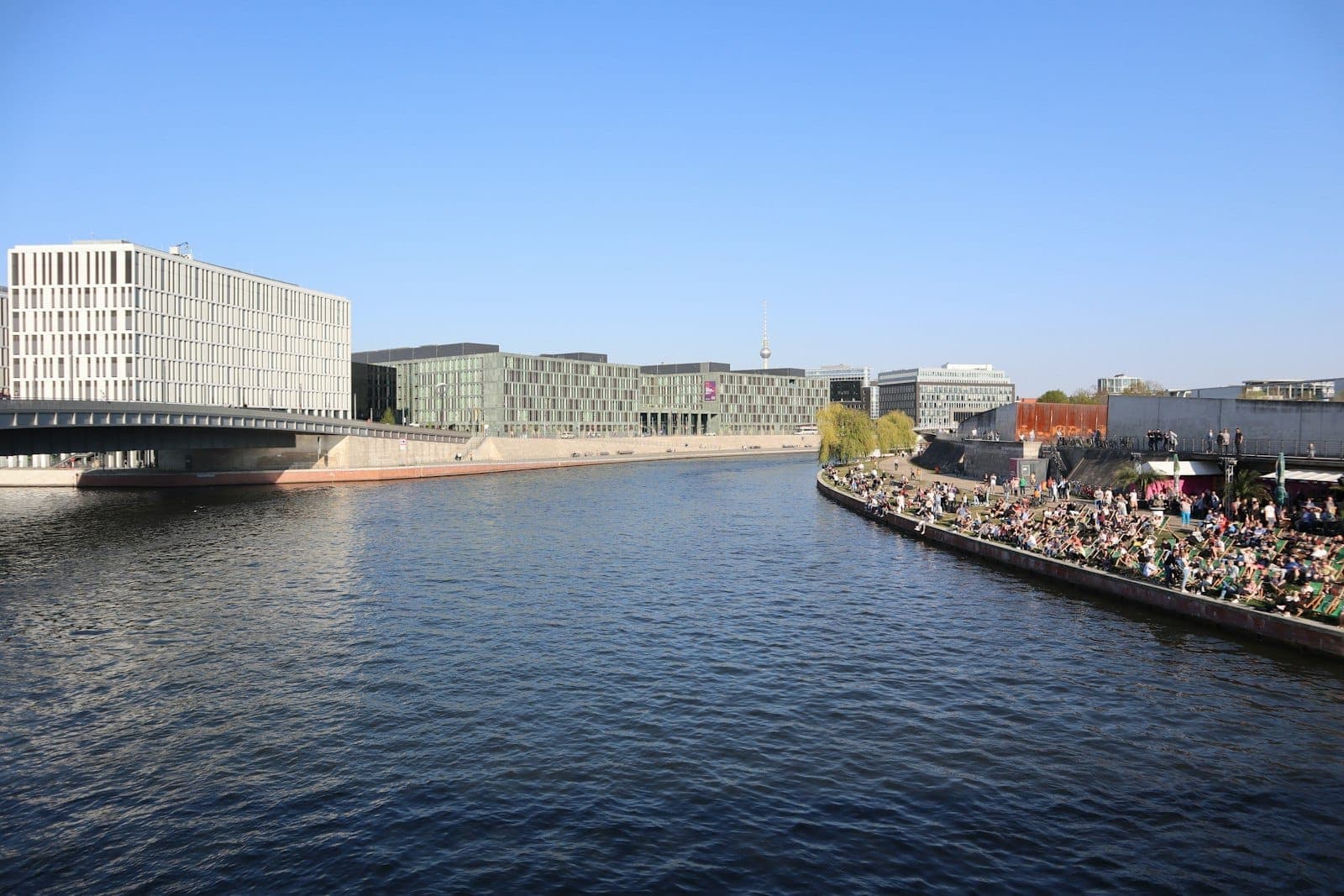 Vista de una concurrida orilla del río Berlín en verano con multitudes relajándose junto al Spree, edificios modernos y un cielo azul despejado.