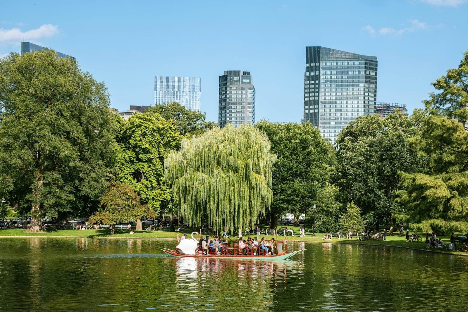 Boston Public Garden au printemps avec un bateau-cygne sur la lagune, des arbres luxuriants et la skyline de la ville en arrière-plan sous un ciel dégagé.