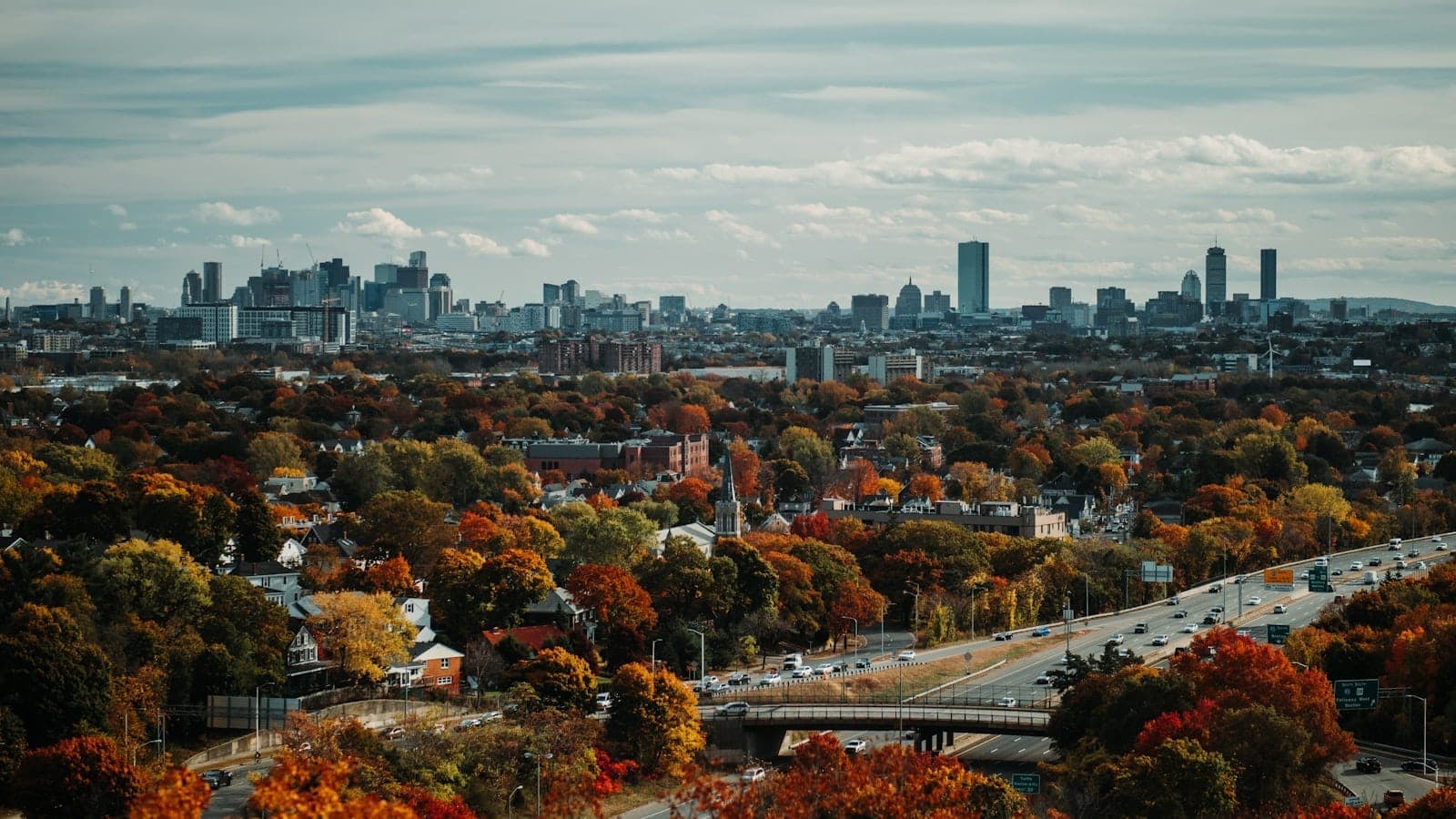 Une vue panoramique sur la skyline de Boston au loin au-dessus d'un vaste paysage d'arbres aux couleurs d'automne vibrantes sous un ciel nuageux