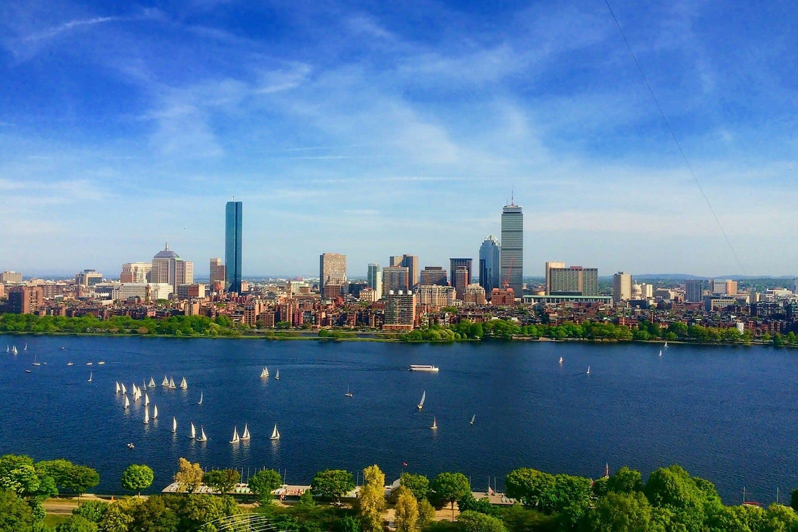 Vue diurne et animée du skyline de Boston avec des voiliers sur la Charles River, des arbres verdoyants au premier plan et un ciel bleu éclatant.