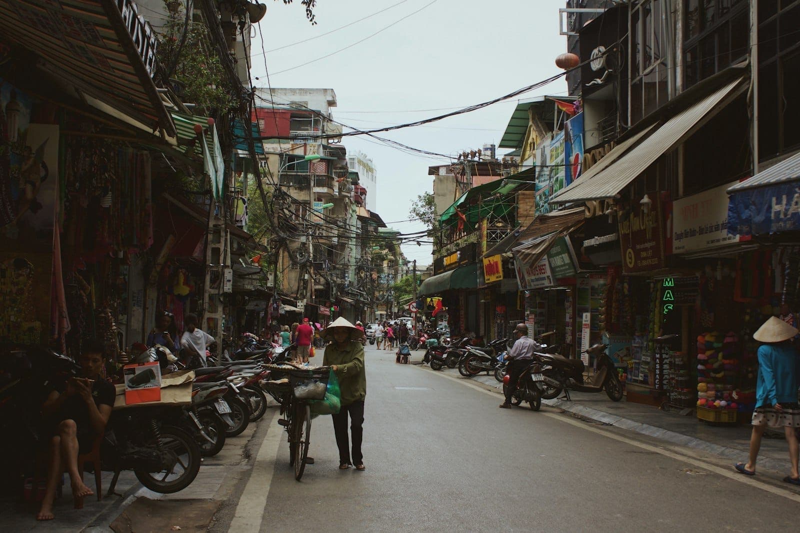 Bustling street scene in Hanoi old quarter with pedestrians, scooters, narrow buildings, and open shopfronts under daylight.