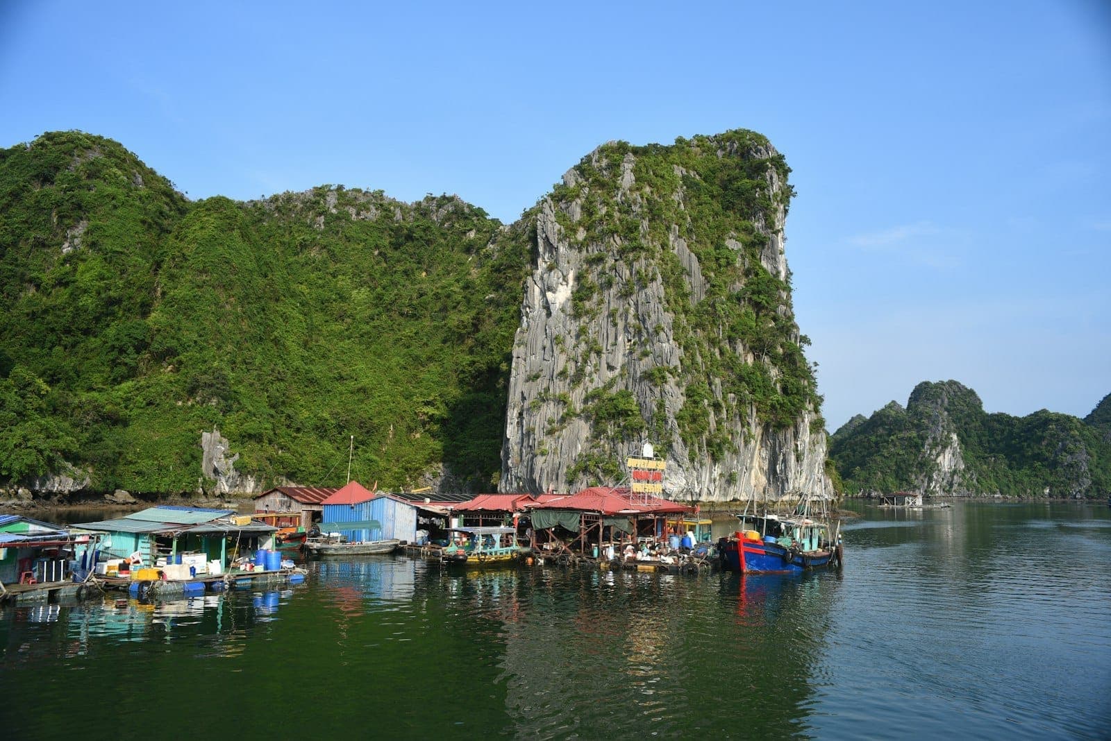 Floating fishing village with colorful buildings and boats set against dramatic limestone karsts and calm water under a clear blue sky.