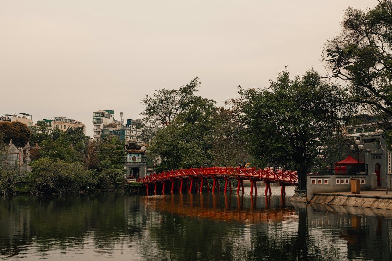 Red wooden bridge across Hoan Kiem Lake with trees and Hanoi skyline in the background under overcast sky.