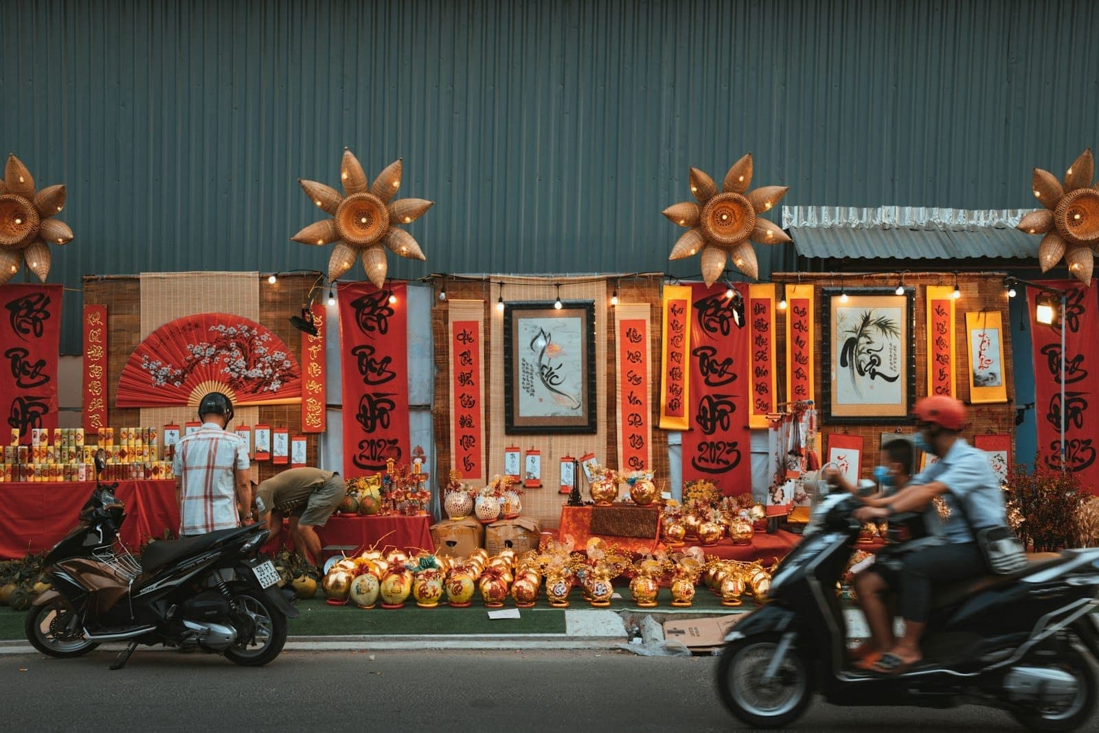 Kalye na may dekorasyon na pulang mga banner, gintong ornamento, at mga bagay na may temang Tết, na may mga scooter na dumadaan sa harap.