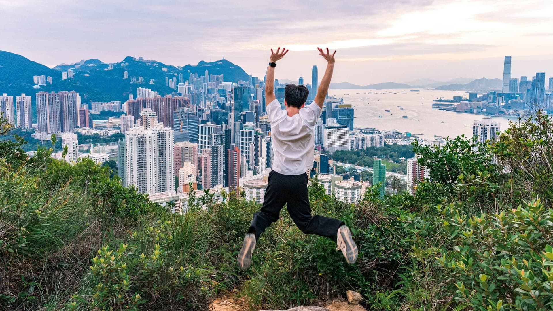 Person jumping on a hillside overlooking Hong Kong skyline and Victoria Harbour with skyscrapers, water, and surrounding hills