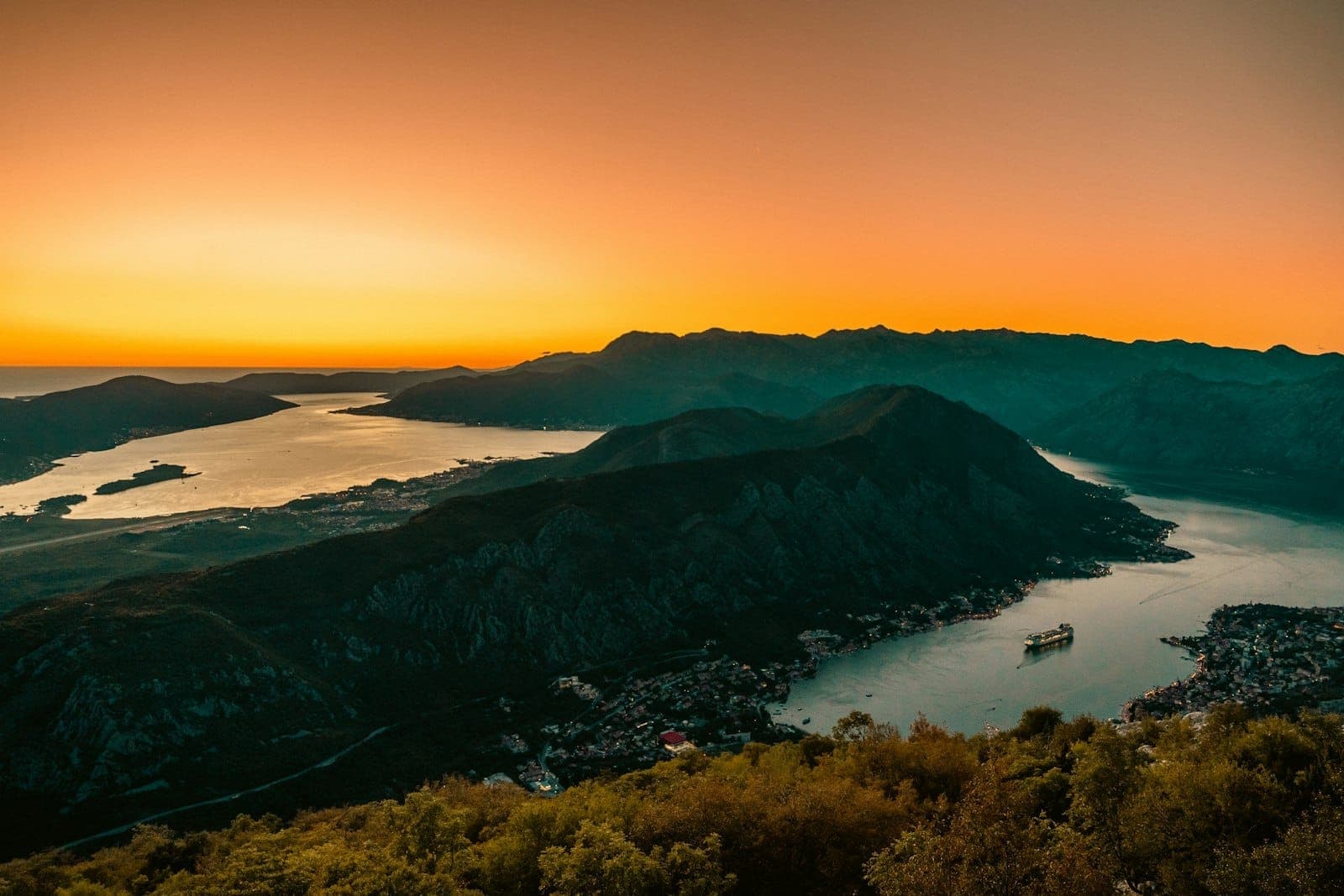 Vue dramatique du coucher de soleil sur la Baie de Kotor avec une lumière dorée d'automne et un bateau de croisière sur l'eau, entourée de montagnes.