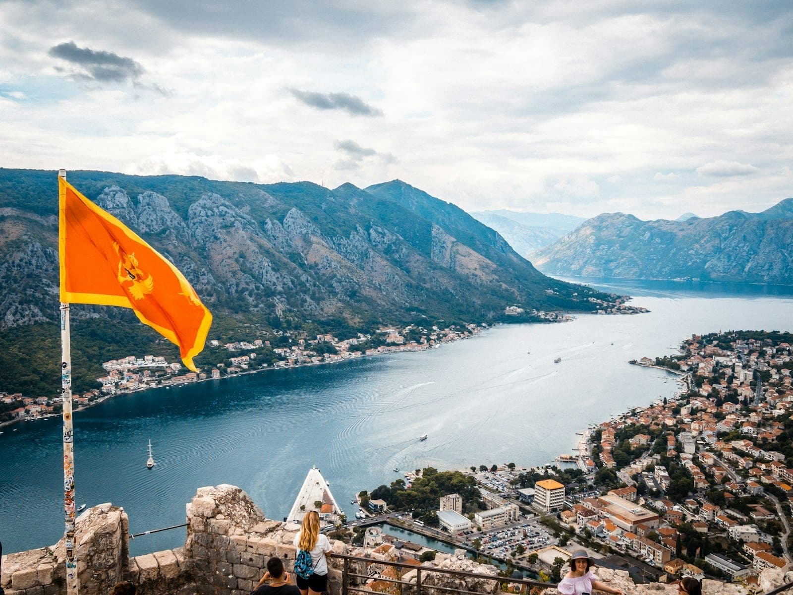Vue panoramique de la Baie de Kotor avec des montagnes, des bâtiments épars et le drapeau monténégrin au premier plan sous un ciel nuageux.