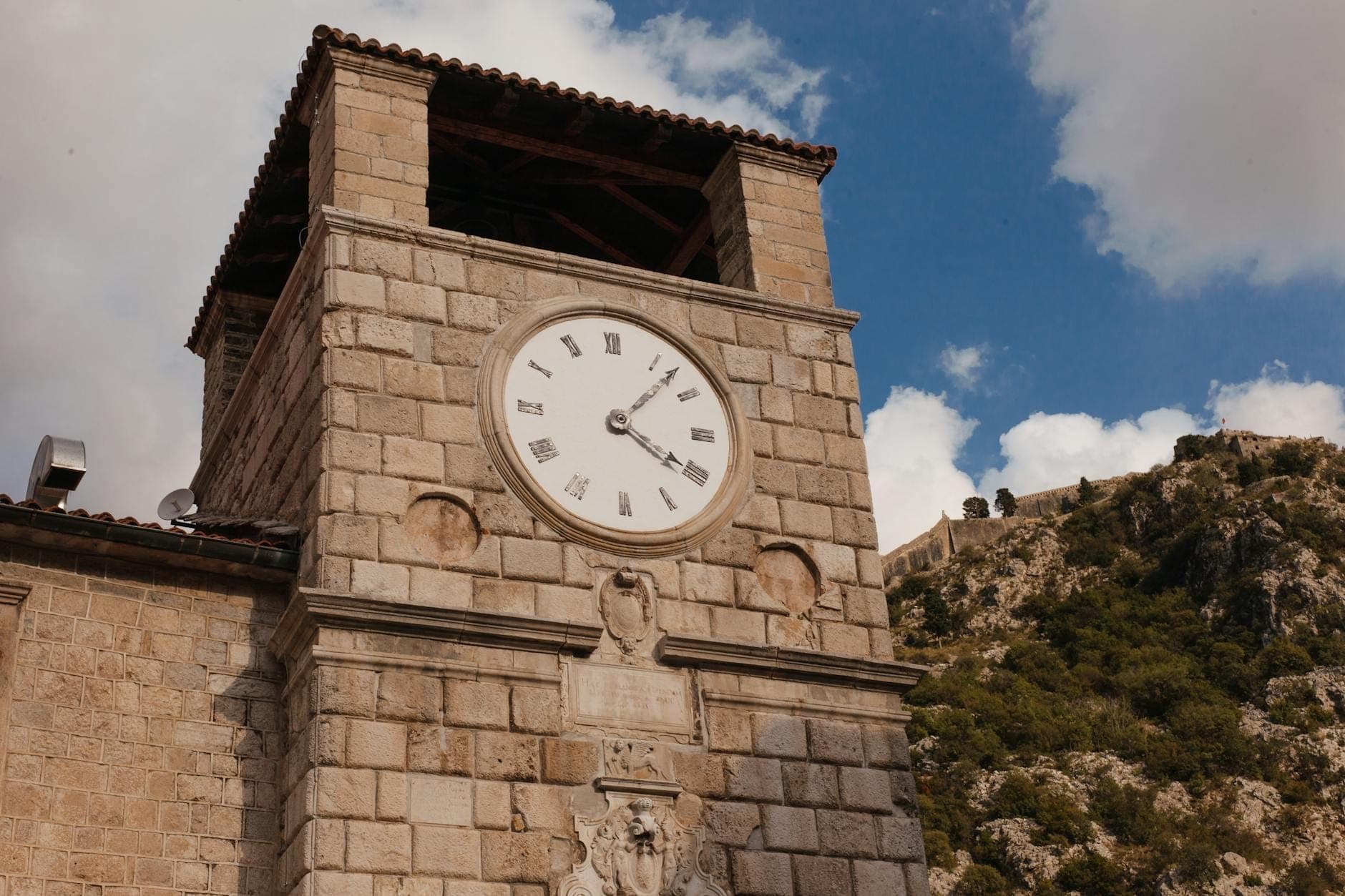 Tour de l'horloge en pierre dans la vieille ville de Kotor avec des chiffres romains et un ciel bleu, avec des montagnes et une forteresse visibles en arrière-plan.