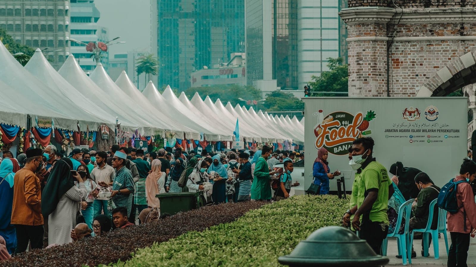 Grande folla che partecipa a un festival gastronomico sotto tende bianche nel centro di Kuala Lumpur, con grattacieli e vegetazione sullo sfondo.