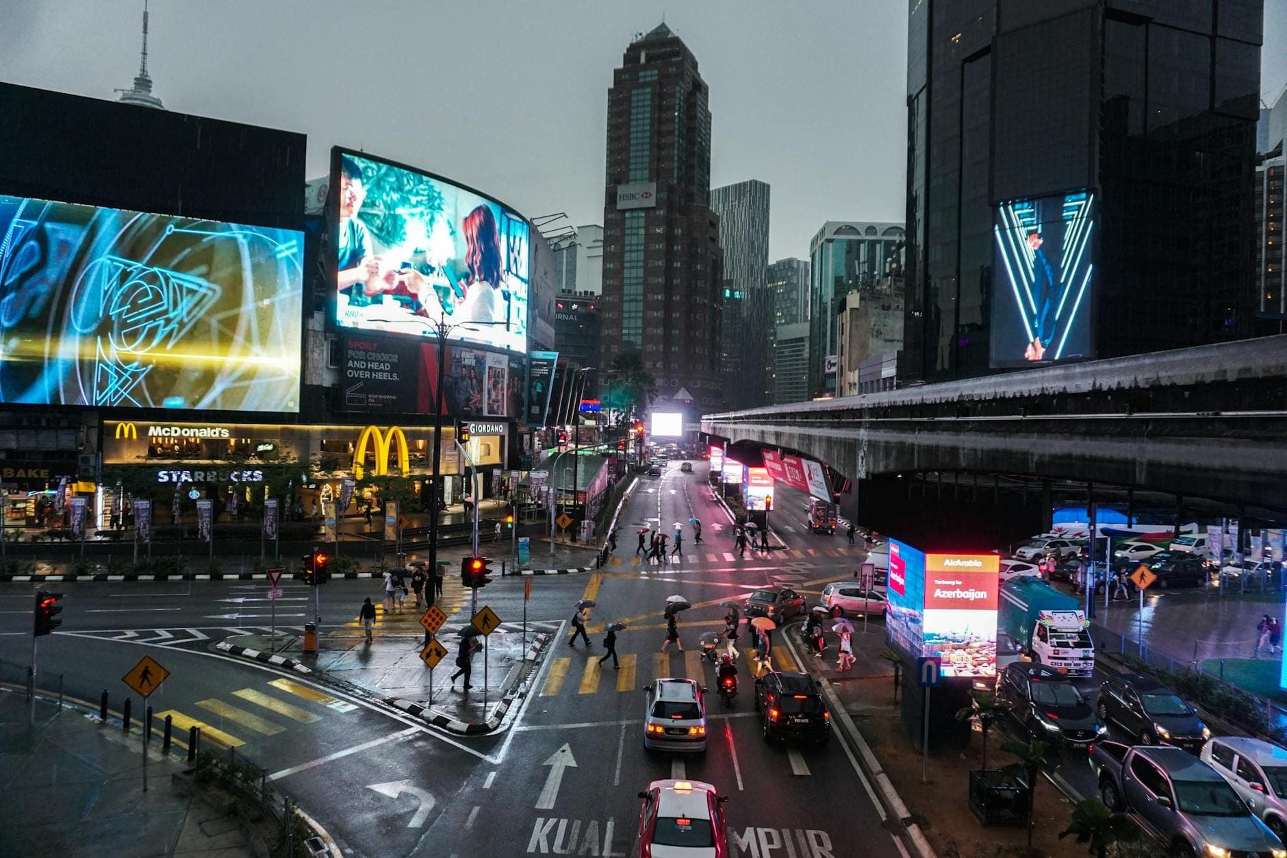Serata piovosa all'incrocio di Bukit Bintang a Kuala Lumpur, con cartelloni luminosi, traffico intenso, pedoni e iconiche insegne di fast food.