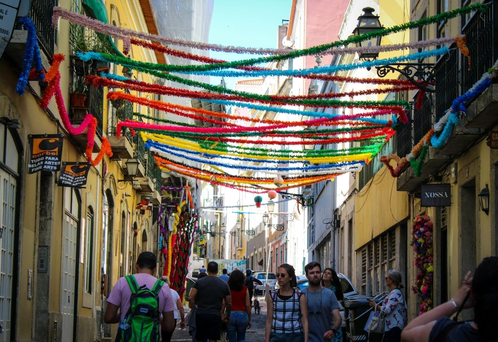 Uma movimentada rua de Lisboa decorada com serpentinas coloridas de festa e pessoas caminhando, criando uma atmosfera animada e festiva.