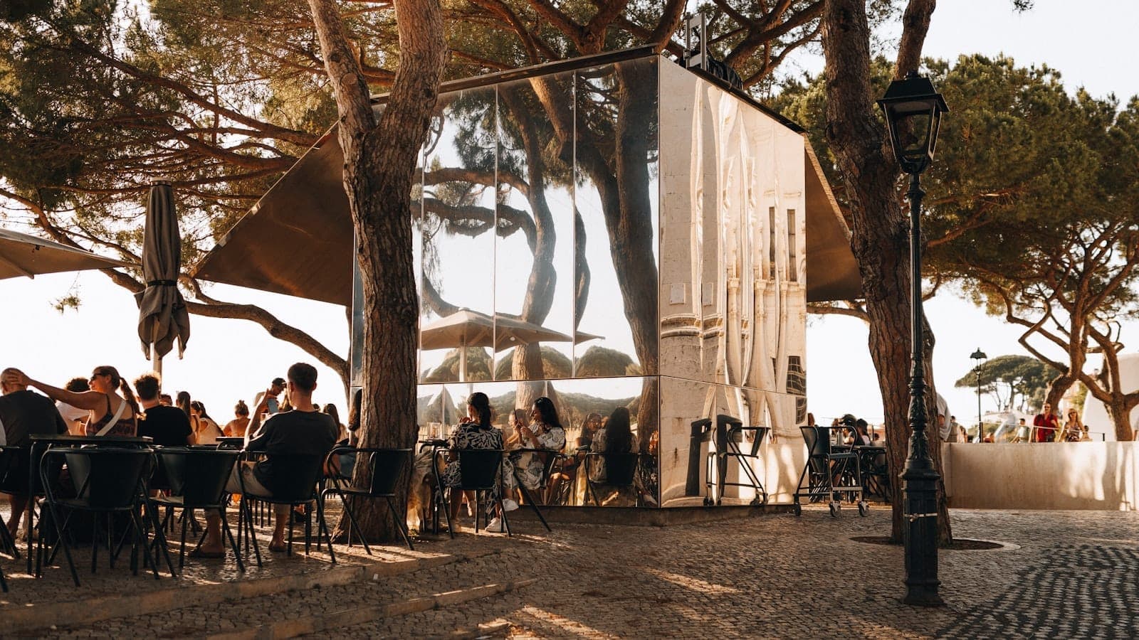 Café ao ar livre em Lisboa sob árvores, com pessoas sentadas em mesas e uma estrutura artística refletora à luz do inverno.