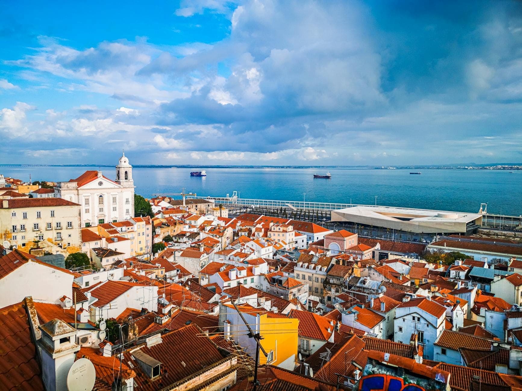 Vista panorâmica de Lisboa com telhados de terracota, edifícios brancos e o oceano Atlântico sob um céu parcialmente nublado.