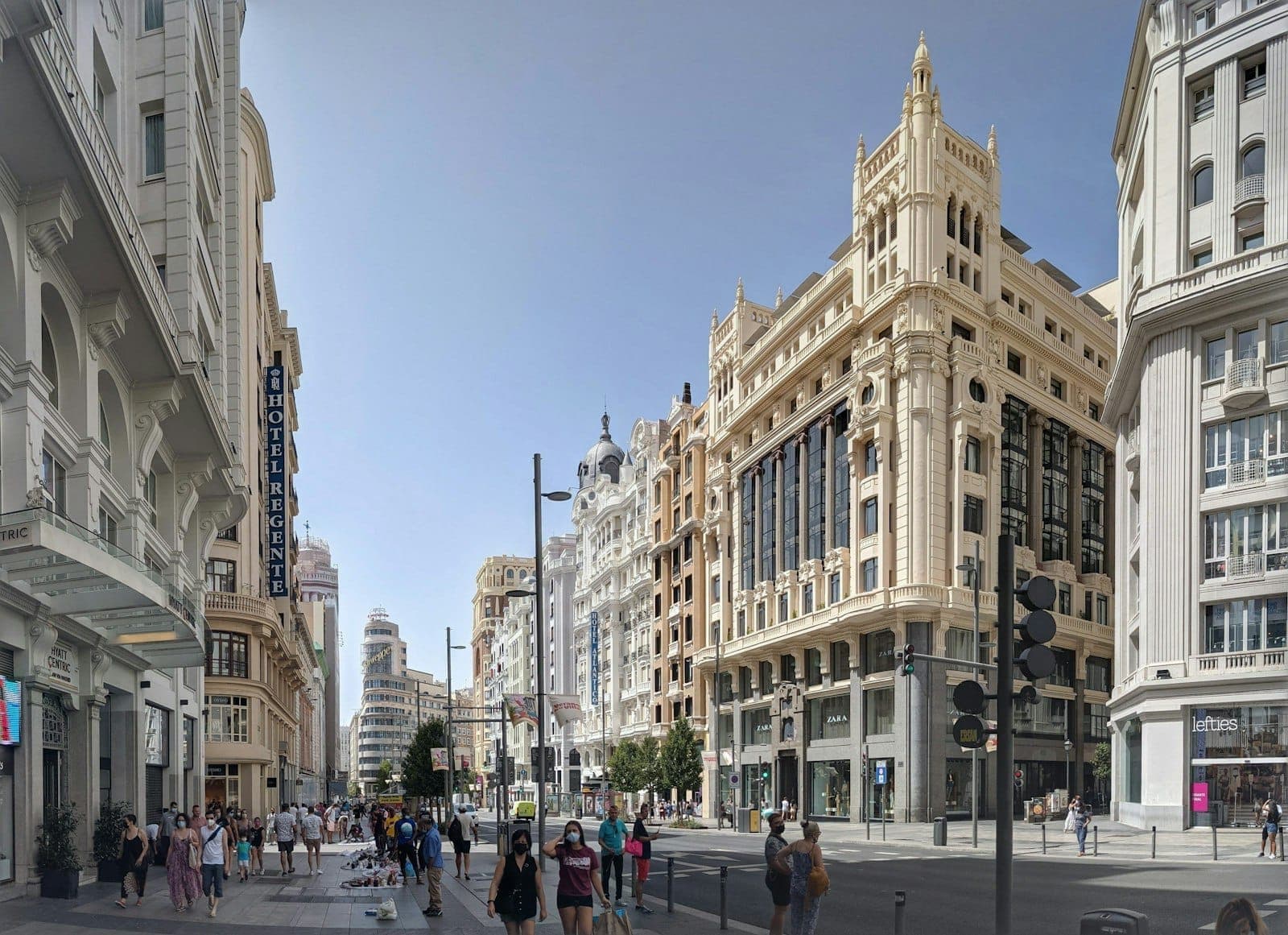 Madrid'de güneşli, kalabalık bir sokak; açık gökyüzünün altında yürüyen insanlar ve klasik şehir mimarisiyle şehrin canlı yaz atmosferini gözler önüne seriyor.