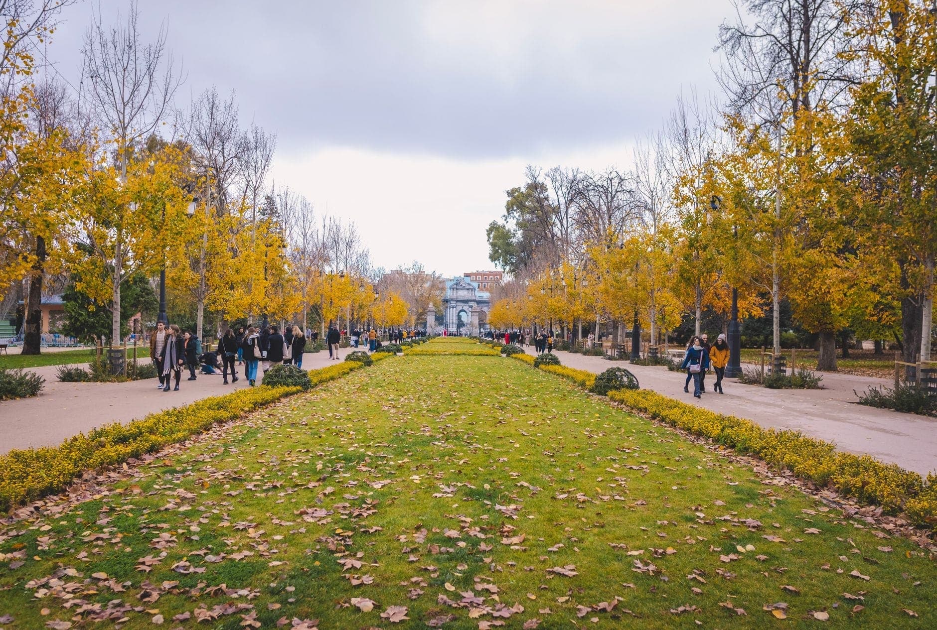 Madrid'deki Parque del Retiro'da sonbahar yaprakları ve saçılmış yapraklarla kaplı ağaçlıklı bulvar, yürüyen insanlar ve bulutlu gökyüzü.