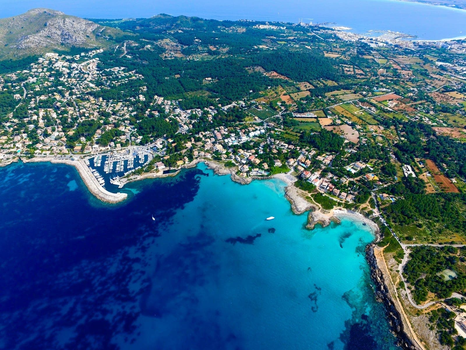 Vista aerea della costa di Mallorca con acque turchesi, marina, paesaggio verde ed edifici sparsi sotto un cielo soleggiato.