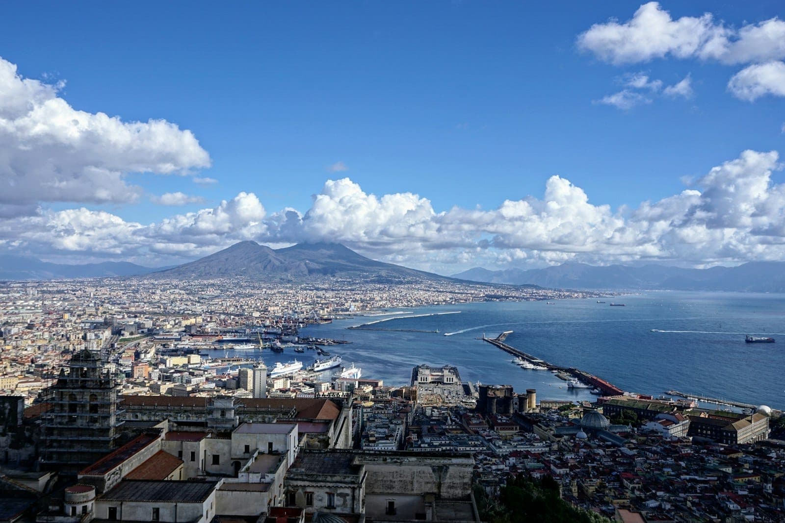 Vista panorâmica de Nápoles com o litoral, os prédios e o Monte Vesúvio ao fundo sob um céu azul com nuvens.
