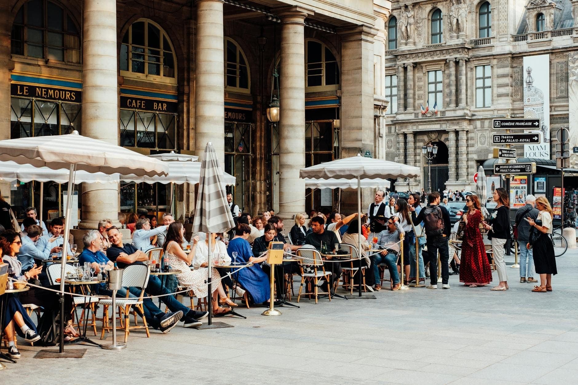 Оживлённая сцена в парижском уличном кафе: толпы людей обедают и общаются под зонтиками рядом с историческими зданиями в солнечный день.