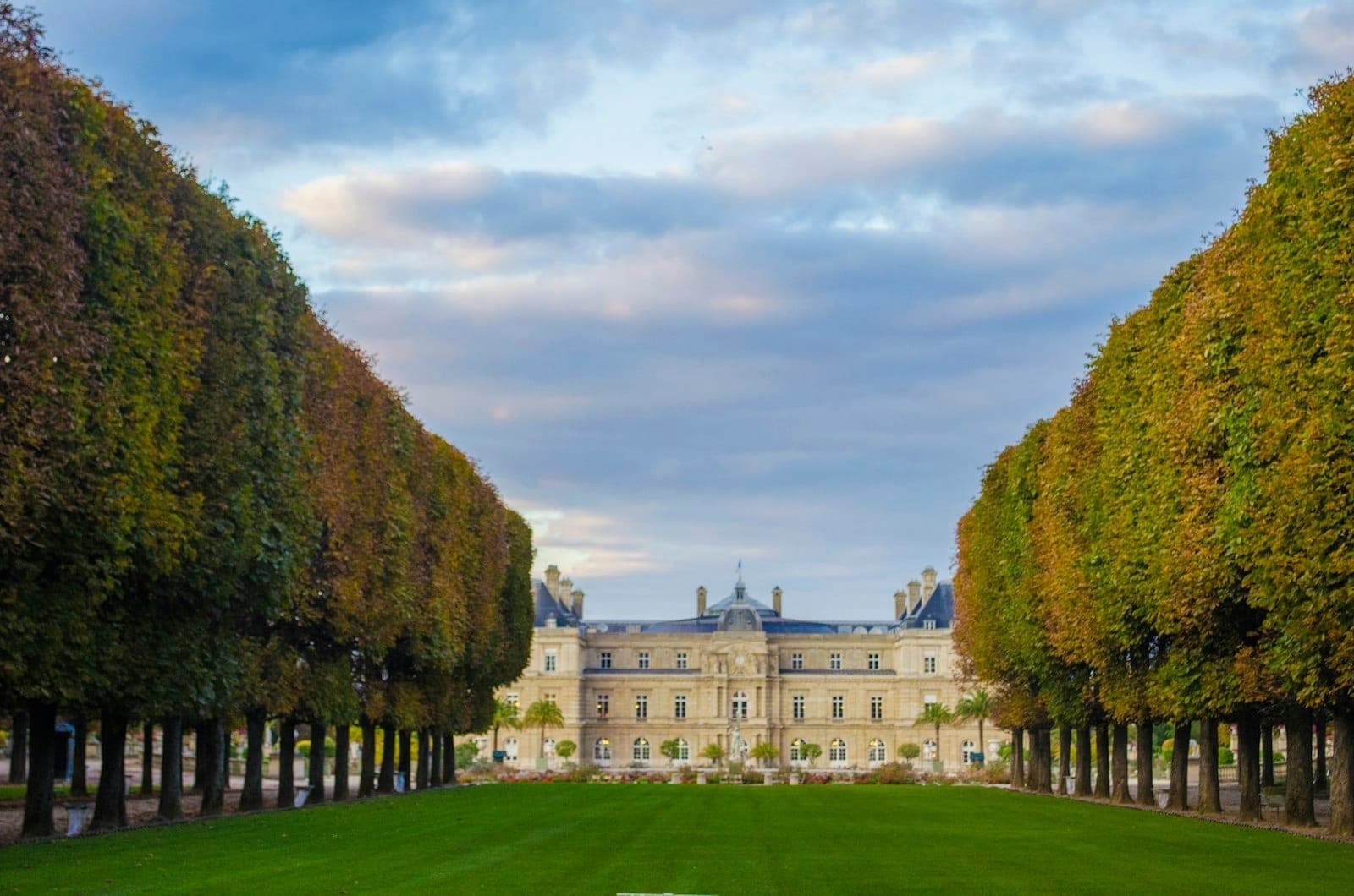 Элегантные ряды деревьев с осенней листвой обрамляют лужайку, ведущую к Palais du Luxembourg в Париже, на фоне частично облачного неба.