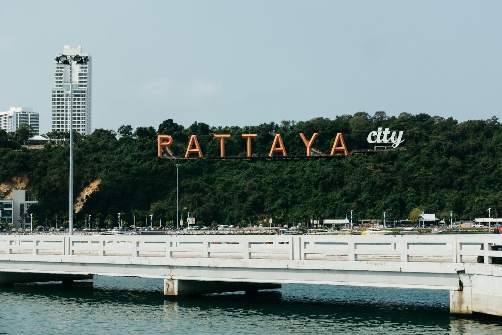Large orange Pattaya City sign on a green hillside, white high-rise buildings in the background, with a bridge crossing over calm water in the foreground.