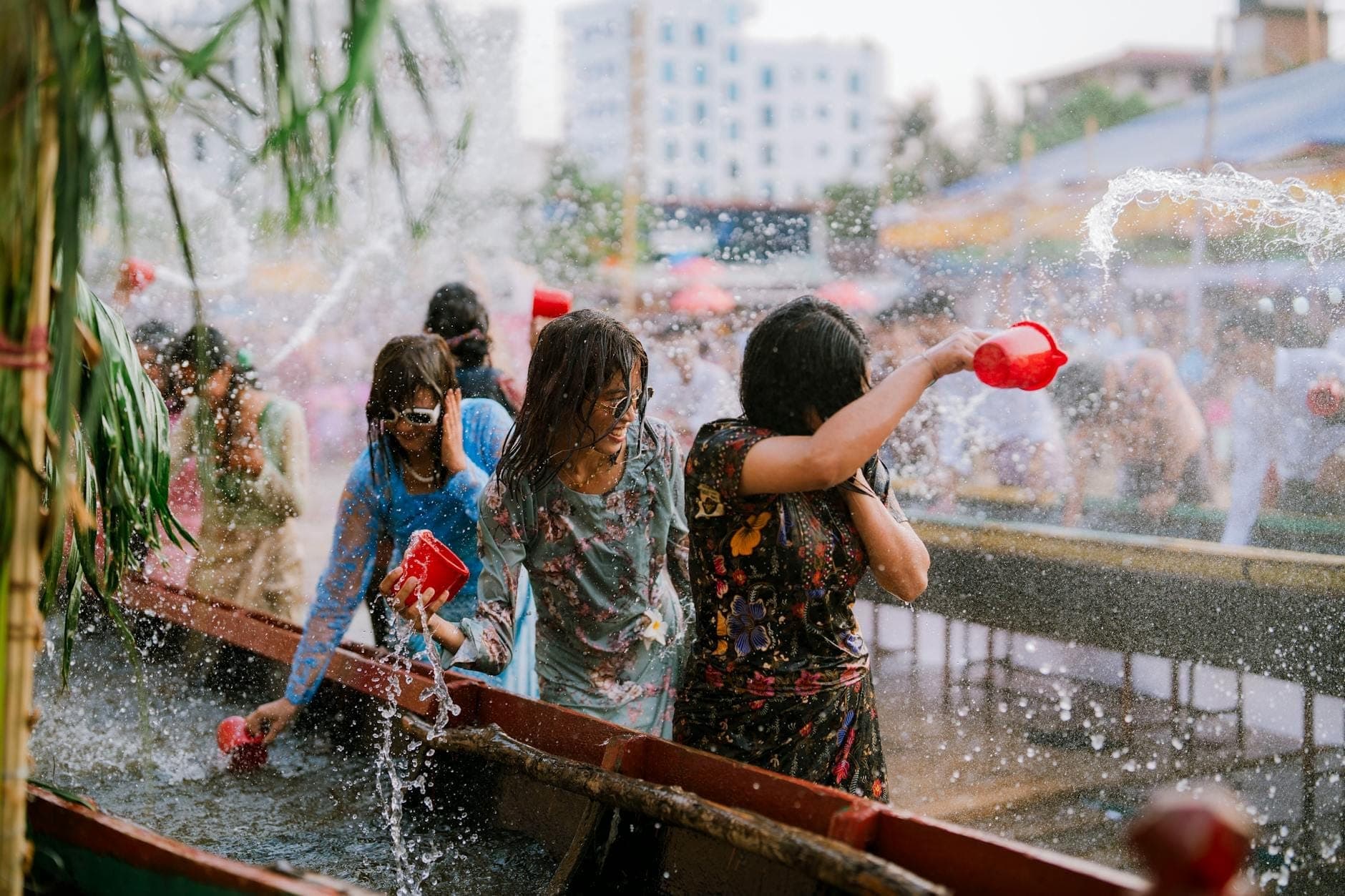 Những người phụ nữ đang vui chơi với nước và té nước vào nhau tại một sự kiện lễ hội Songkran ngoài trời ở Pattaya, với các thùng chứa nước và các tòa nhà thành phố hiện ra phía sau.