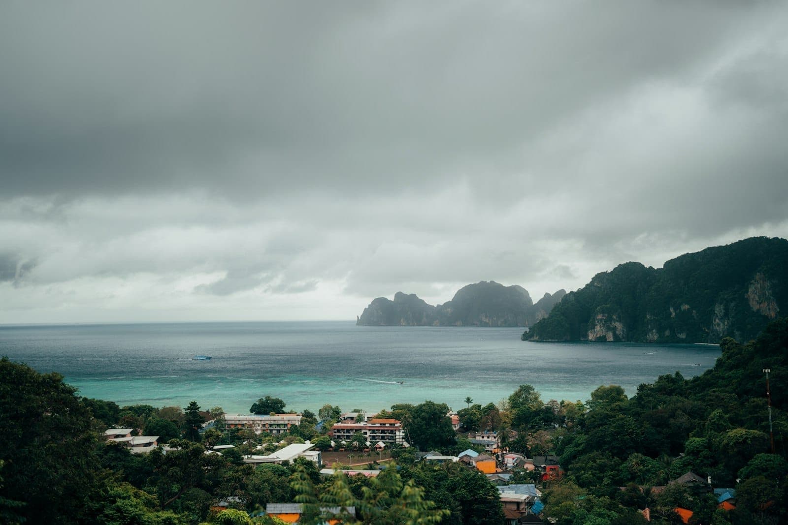 Dramatikong makulimlim na langit sa ibabaw ng isang bayang baybayin sa Phuket na may berdeng mga burol at turkesang tubig, na nagpapahiwatig ng panahon ng monsoon.