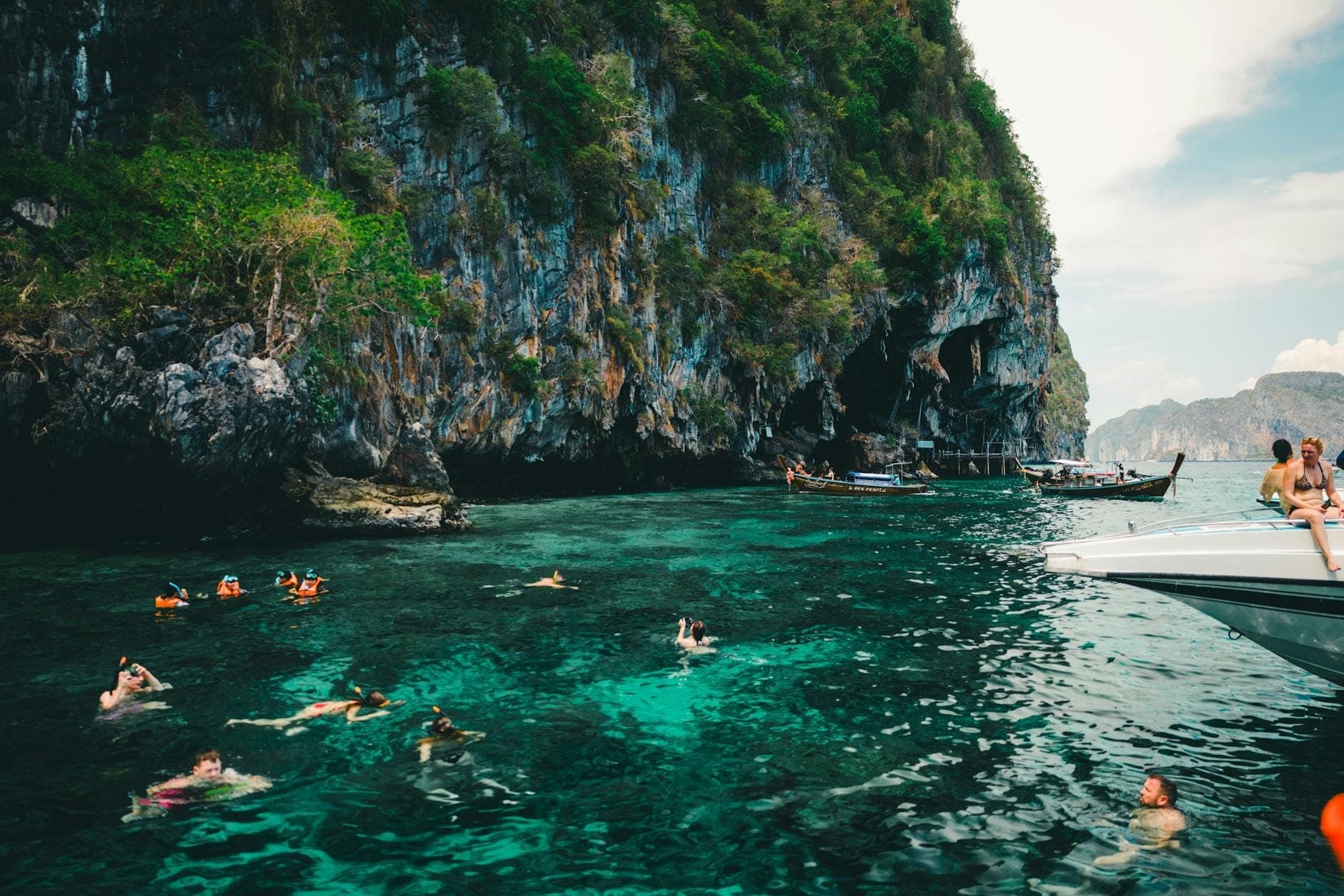 Mga turista na lumalangoy at nag-snosnorkeling sa malinaw na tubig na turkesa malapit sa mga dramatikong limestone cliffs, na may mga bangka at malusog na halaman sa likod, katangian ng mga adventure activities sa Phuket.