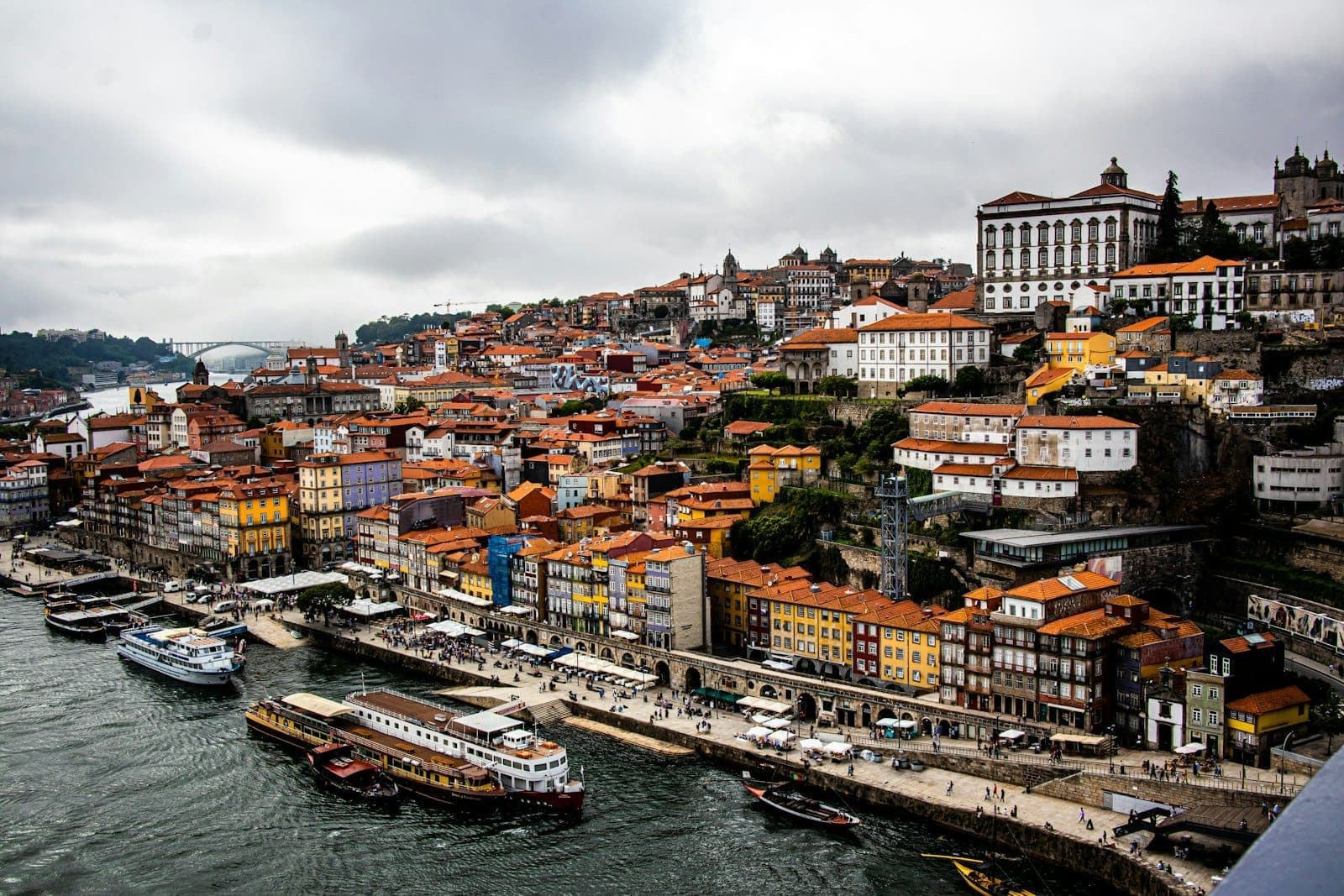 Animado paseo fluvial de Porto con coloridos edificios históricos, barcos y multitudes a lo largo del malecón en un día nublado.