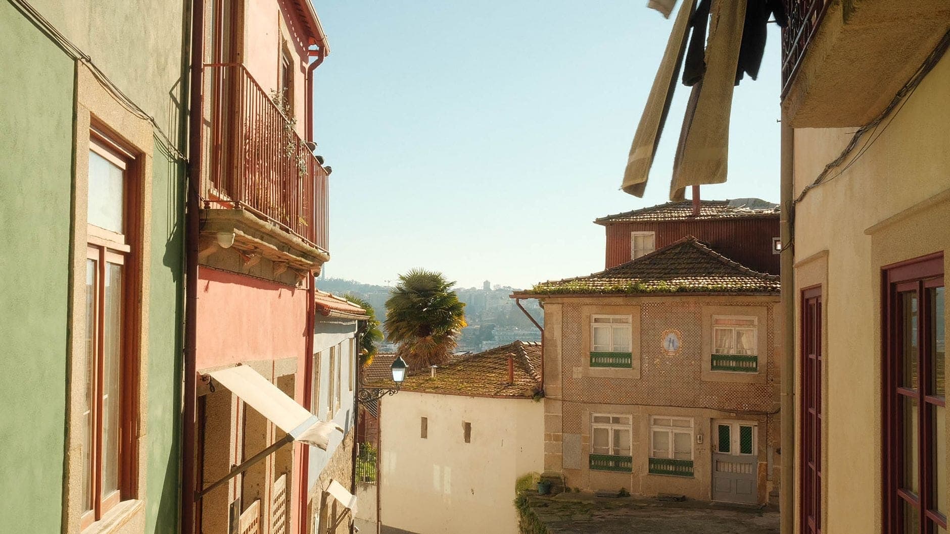 Una tranquila calle soleada de Porto bordeada de casas coloridas y balcones, ropa tendida afuera y un atisbo del río Duero a lo lejos.