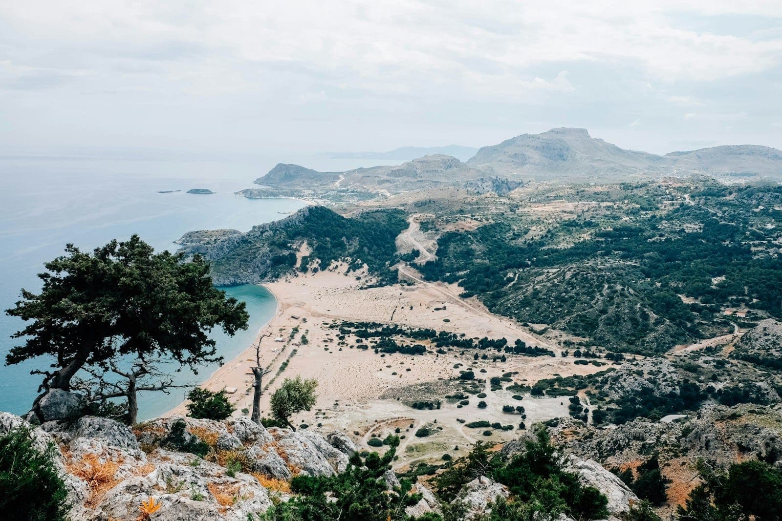 Panoramiczny widok na wybrzeże Rodos z piaszczystymi plażami, krystalicznie błękitnym morzem i skalistymi górami pod zachmurzonym niebem.